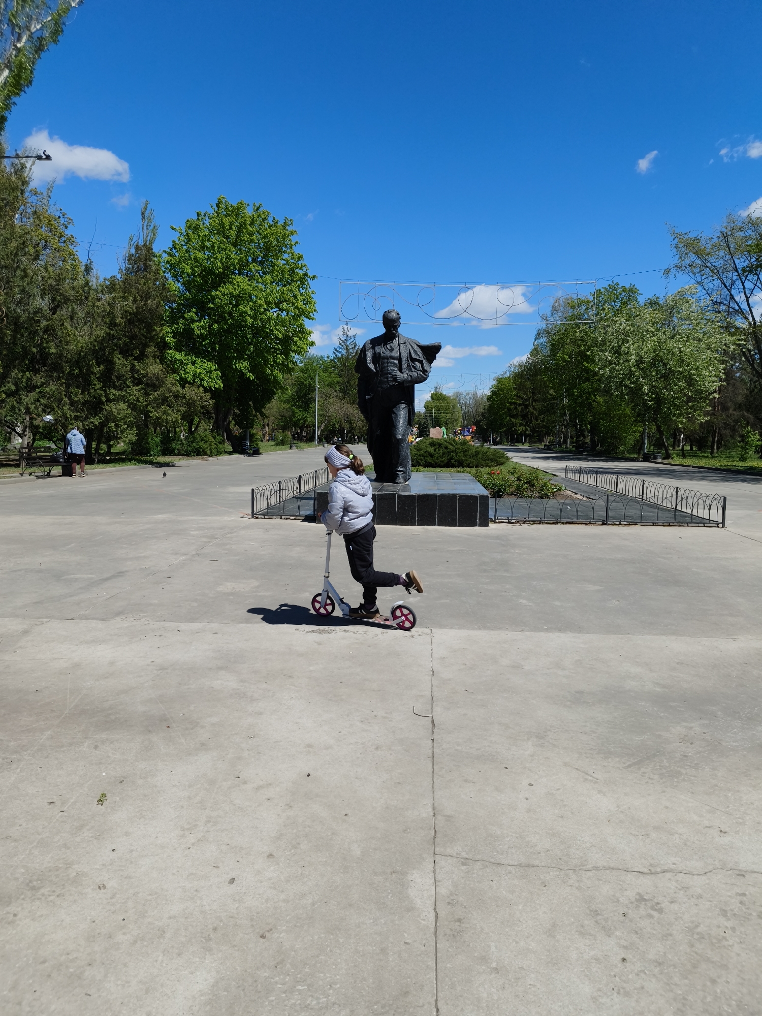 This outdoor image captures a vibrant public park or square in Kryvyi Rih, Ukraine, on a bright, clear day. The sky is a brilliant blue with scattered white clouds, indicating pleasant weather. Sunlight casts distinct shadows, suggesting it is likely mid-morning or early afternoon.
The central focus is a child or young person, seen from behind, actively riding a white and pink scooter across a wide, light-colored concrete expanse. They are dressed in a light grey jacket, dark trousers, and a grey headband, pushing off with one foot.
Dominating the mid-ground is a large, dark-colored statue of a male figure, standing on a polished black marble base. The figure, dressed in what appears to be a suit, stands with one arm slightly raised, characteristic of historical monuments often found in post-Soviet cities. A low metal fence surrounds the statue's base. Above the statue, a white, decorative metal structure with looping patterns stretches across the sky.
The setting is lush with greenery. Various trees with full, bright green foliage frame the square, providing shade. In the left background, another person in a light blue jacket stands or sits near a bench, further indicating the park setting. A small, dark bird is visible on the concrete ground between the scooter rider and the person on the bench. A paved path curves away to the right, disappearing into more trees.
There is no visible text in the image. The scene conveys a sense of peaceful leisure and outdoor activity within a historic urban park landscape.