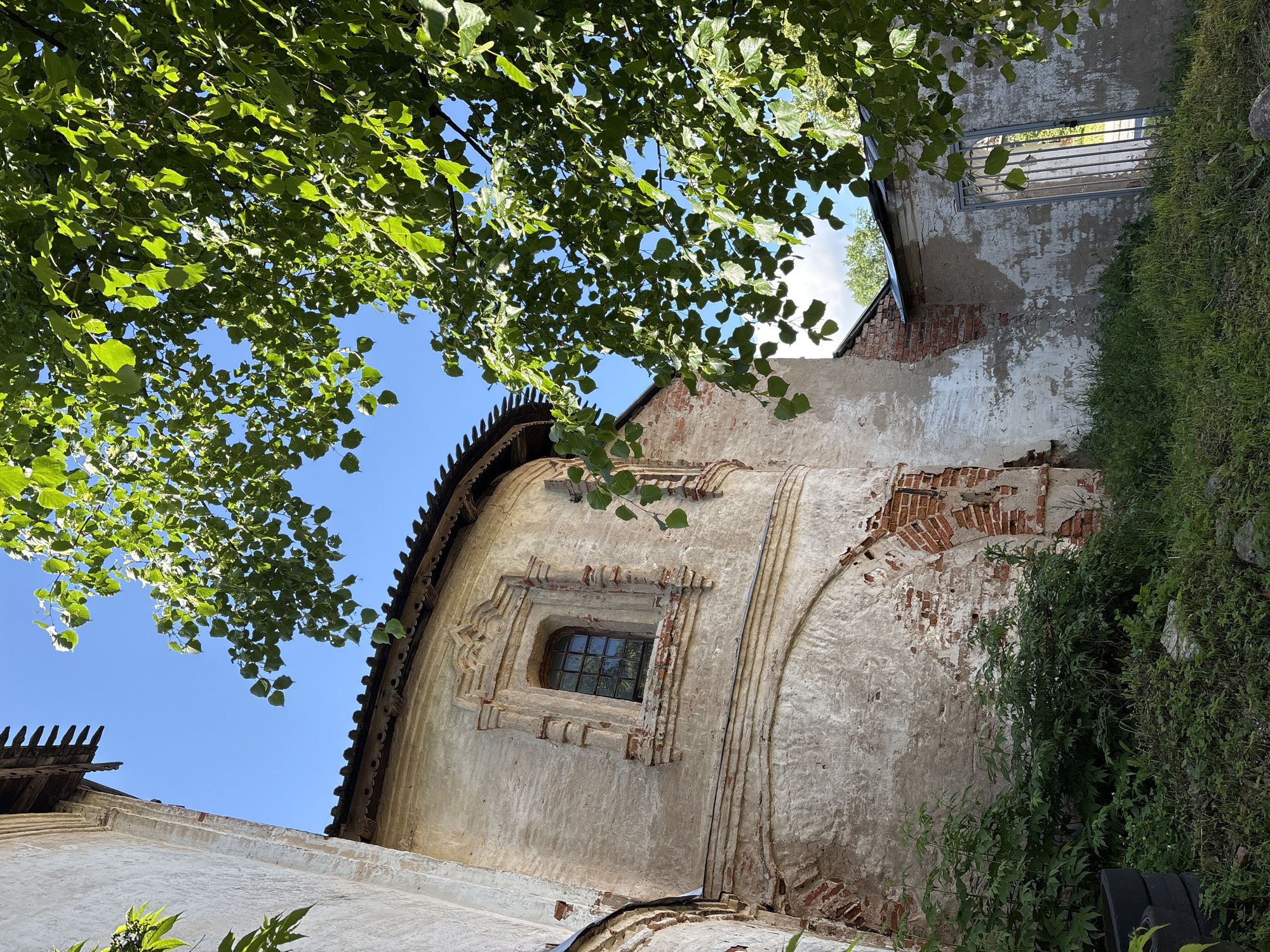 The image shows a partial view of an old, weathered building with peeling plaster and exposed brickwork. The architecture suggests a historical structure, possibly religious in nature, given the ornate window and curved wall. Lush green foliage from a tree partially obscures the upper right portion of the frame, with its leaves contrasting against a clear blue sky. The ground is covered in overgrown grass and some dark, possibly discarded, objects are visible on the left. A metal gate is visible in the background on the right. The scene appears to be set outdoors during the daytime, likely in a rural or less-maintained urban area. There are no people or animals present. This scene is likely located in Veliky Novgorod, Russia, a city known for its rich history and numerous ancient buildings.