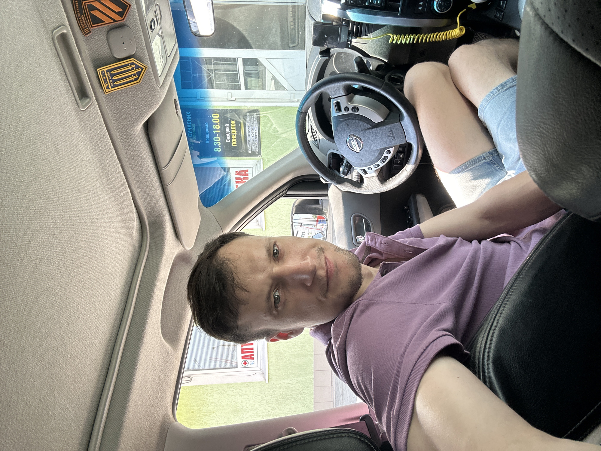 A man is seated in the driver's seat of a car, looking towards the camera. He is wearing a light purple polo shirt and blue denim shorts. The car's interior is visible, including the steering wheel, dashboard, and part of the passenger seat. Outside the car, a building with a lime green facade and a sign with red lettering and a white cross is visible. The image appears to be taken during the daytime, with sunlight entering the car through the windshield and side windows. The location is identified as Kamianets-Podilskyi, Ukraine.