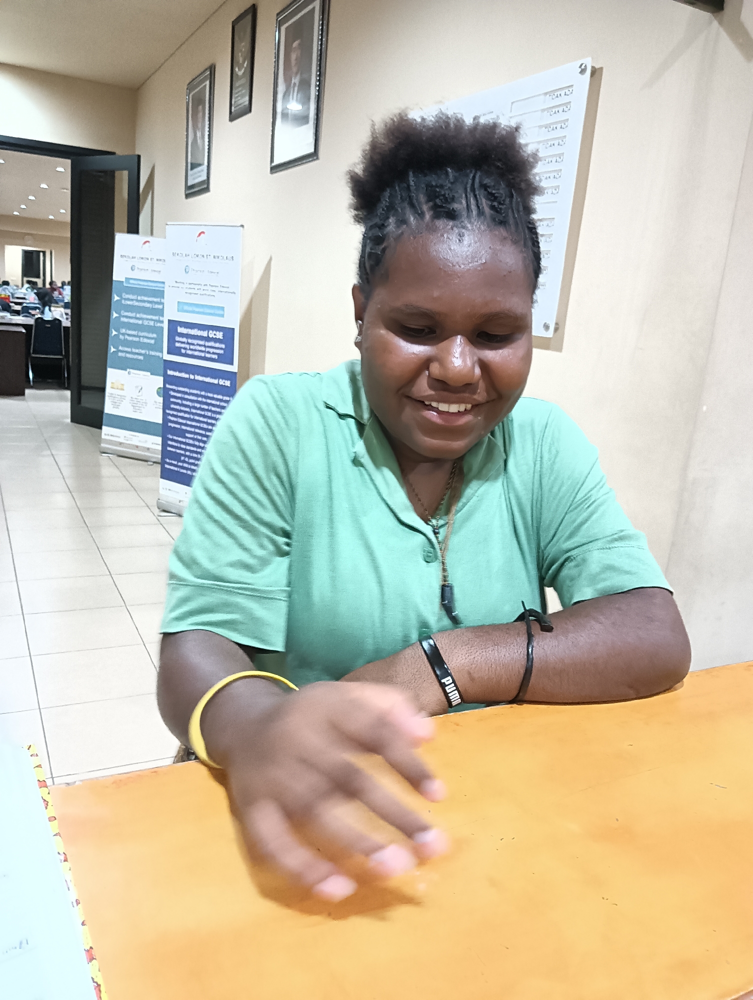 A young person with dark skin and braided hair is seated at an orange table, smiling and looking down. They are wearing a light green collared shirt, a necklace with a dark pendant, a yellow bracelet on their left wrist, and a black bracelet on their right wrist. Their right hand is blurred as it moves across the table. In the background, two banners with text and graphics are visible, one of which promotes "International GCSE." Several framed pictures hang on a beige wall behind the banners. To the left, a doorway leads to a room with tables and chairs, possibly a classroom or meeting space. The flooring is tiled in a beige and white checkerboard pattern. The overall environment appears to be an indoor educational or administrative setting.