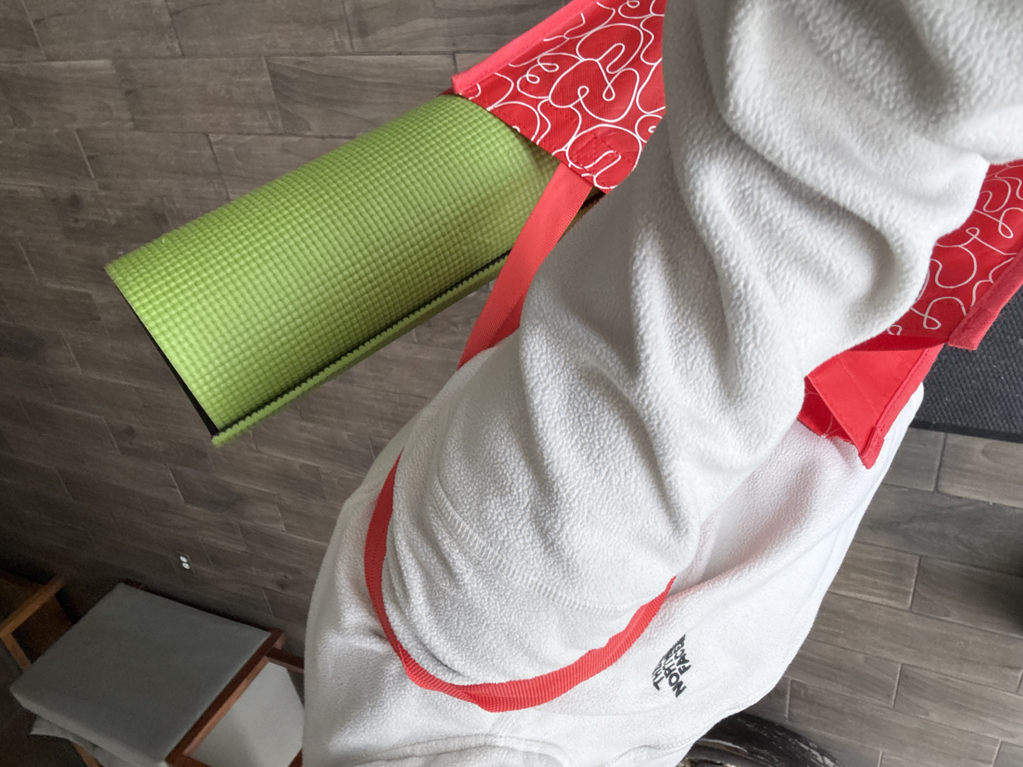 A person wearing a white North Face jacket with red trim and carrying a green yoga mat in a red and white patterned carrier is standing on a floor covered in gray planks. Also visible are a light gray stool with a wooden frame, and a gray yoga mat on the floor. The specific location in Mexico City, Mexico is not discernible from this image.