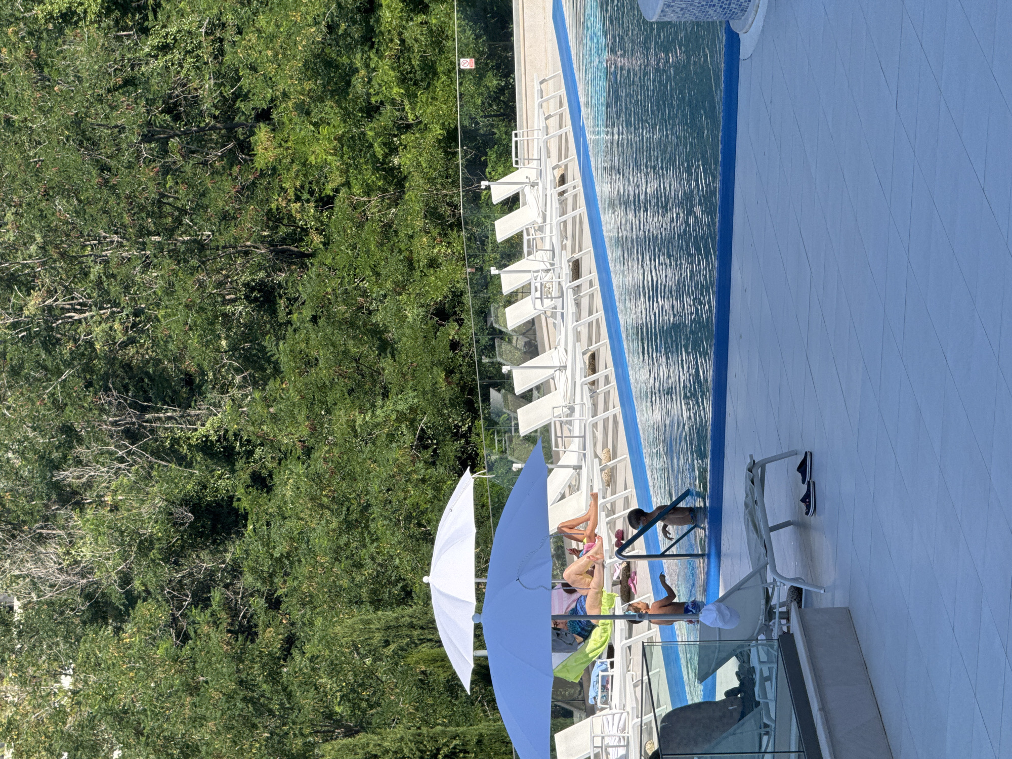 The image shows a swimming pool area at a resort in Boreti, Montenegro. Two young boys are in the pool, one on the left near the steps and the other in the center. A blue umbrella shades a lounge chair where two adults are relaxing, and a white umbrella is positioned behind it. Several white lounge chairs are arranged around the pool. The pool has a blue border and is surrounded by a tiled deck. Beyond the pool area, a dense green forest forms the background. The lighting suggests it is daytime, likely in the morning or afternoon, with clear skies.