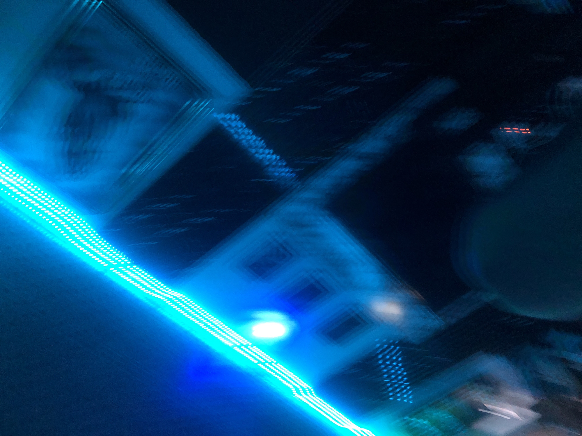 The image shows a diagonal line of white LED lights against a dark blue background. The lights are slightly blurred, suggesting movement or a shallow depth of field. To the right of the lights, there are several out-of-focus geometric shapes and patterns in various shades of blue and black. Some of these shapes appear to be rectangular or square, with visible lines or grids within them. There is a bright white, circular light source located above and to the left of the center of the image, which is also blurred. The overall impression is abstract and impressionistic, with a strong emphasis on light and color. No specific location within a city or any discernible people or actions are present.