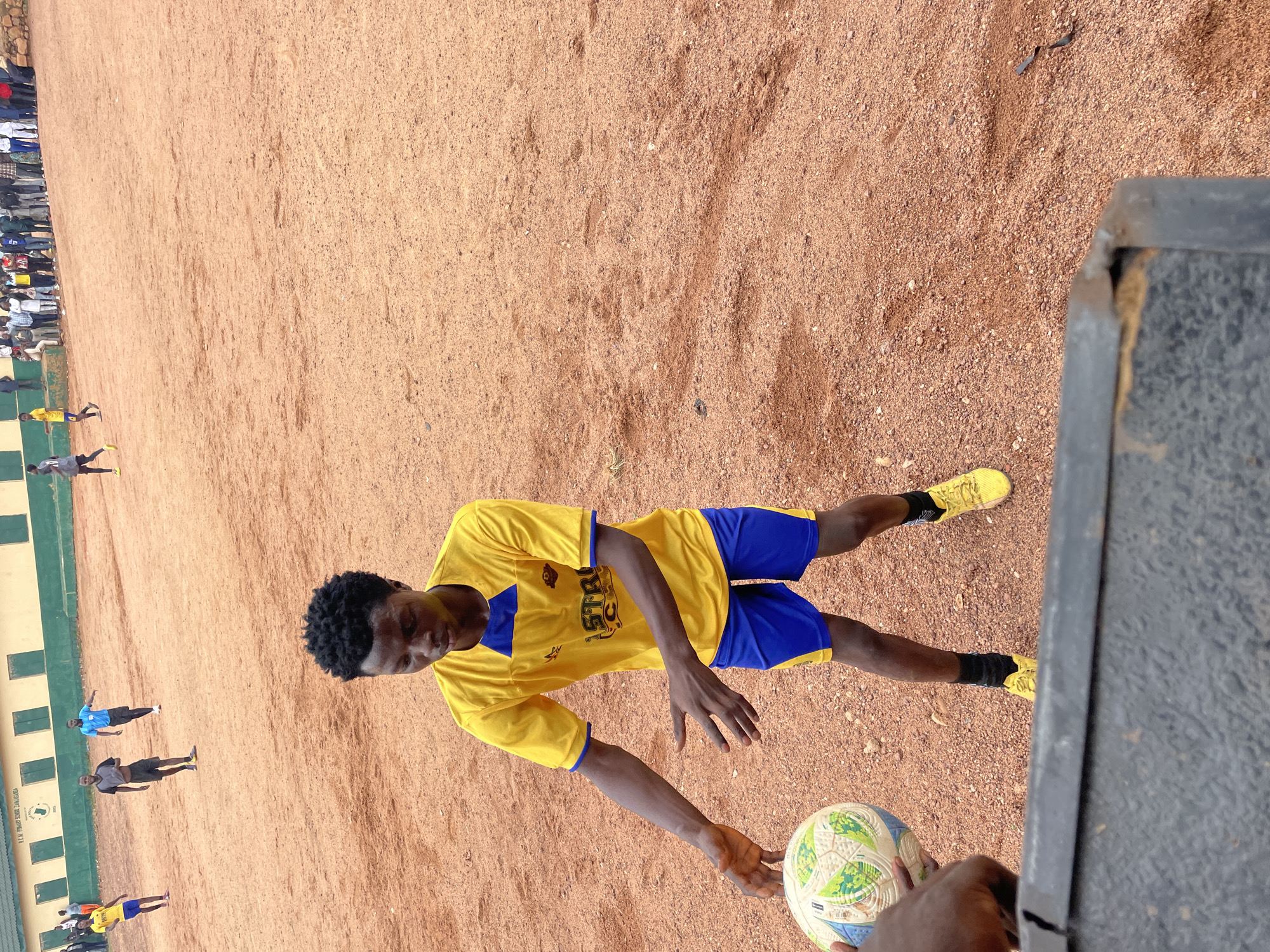A young Black male in a yellow t-shirt and blue shorts with yellow trim is playing soccer on a dirt field. He is looking down at the ball, which is a white, green, and blue soccer ball with a graphic pattern. His arms are outstretched, with his left hand reaching for the ball. Other individuals are visible in the background, some of whom appear to be playing soccer. In the far background, a green building with the words "R.C.M. PRIMARY SCHOOL, ZARIA ROAD, JOS" is visible. The field is a reddish-brown dirt, and there are spectators on a ledge in the background. The scene appears to be a casual soccer game on a school field in Jos, Nigeria.