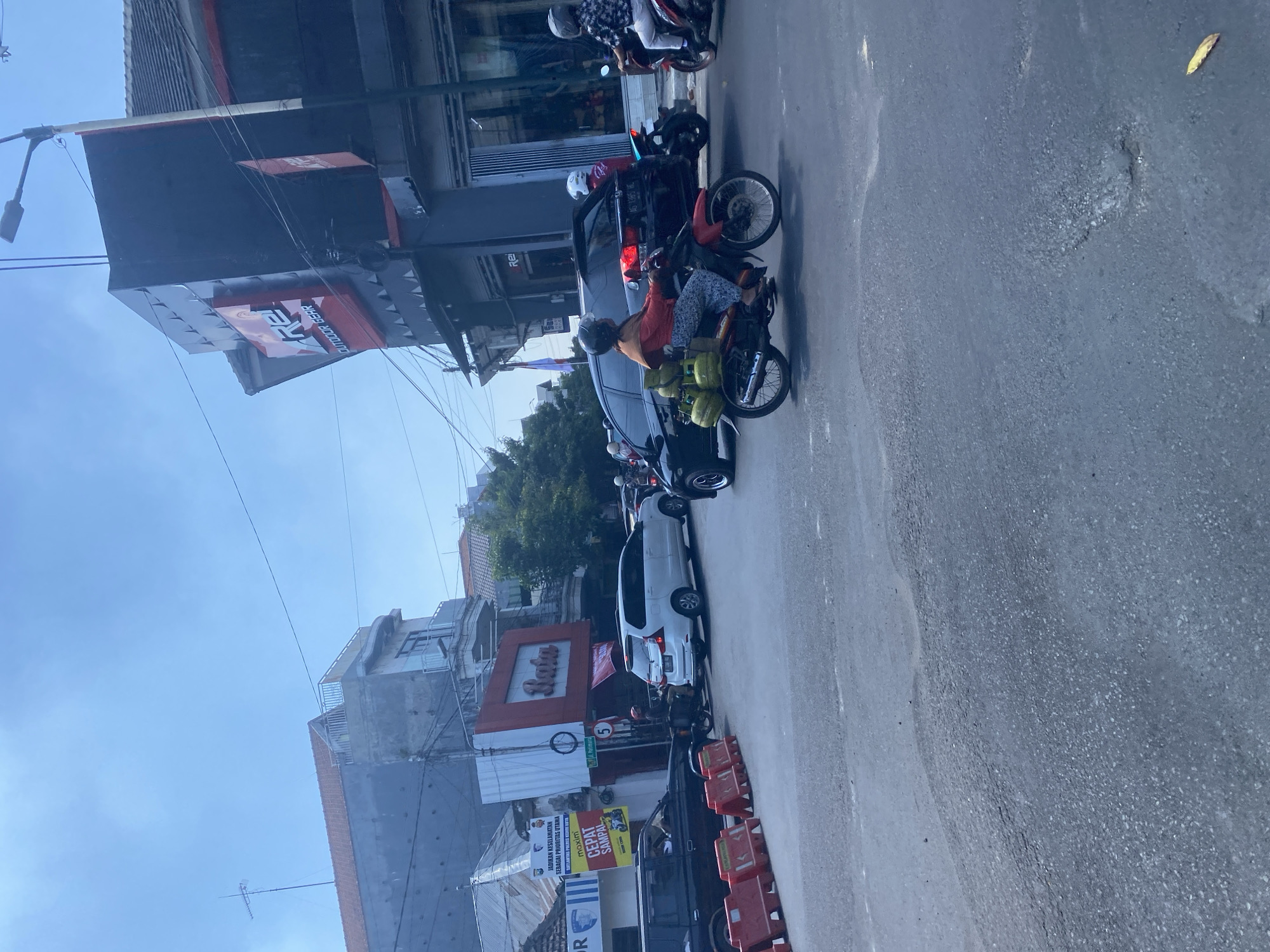 In Kediri City, Indonesia, on a slightly overcast day, a busy intersection is captured. A red motorcycle is ridden by a person wearing an orange headscarf and a patterned blue and white outfit, transporting three green gas cylinders on the rear. The motorcycle is followed by a black car. Further down the road, a white minivan is visible, and beyond that, a building with a prominent red sign that reads "Bata". Another building displays the "REI" logo, indicating an outdoor gear store. The street is paved with asphalt, and some sections show signs of wear and tear, with visible potholes. Red traffic barriers are placed on the side of the road. The sky is a clear blue.