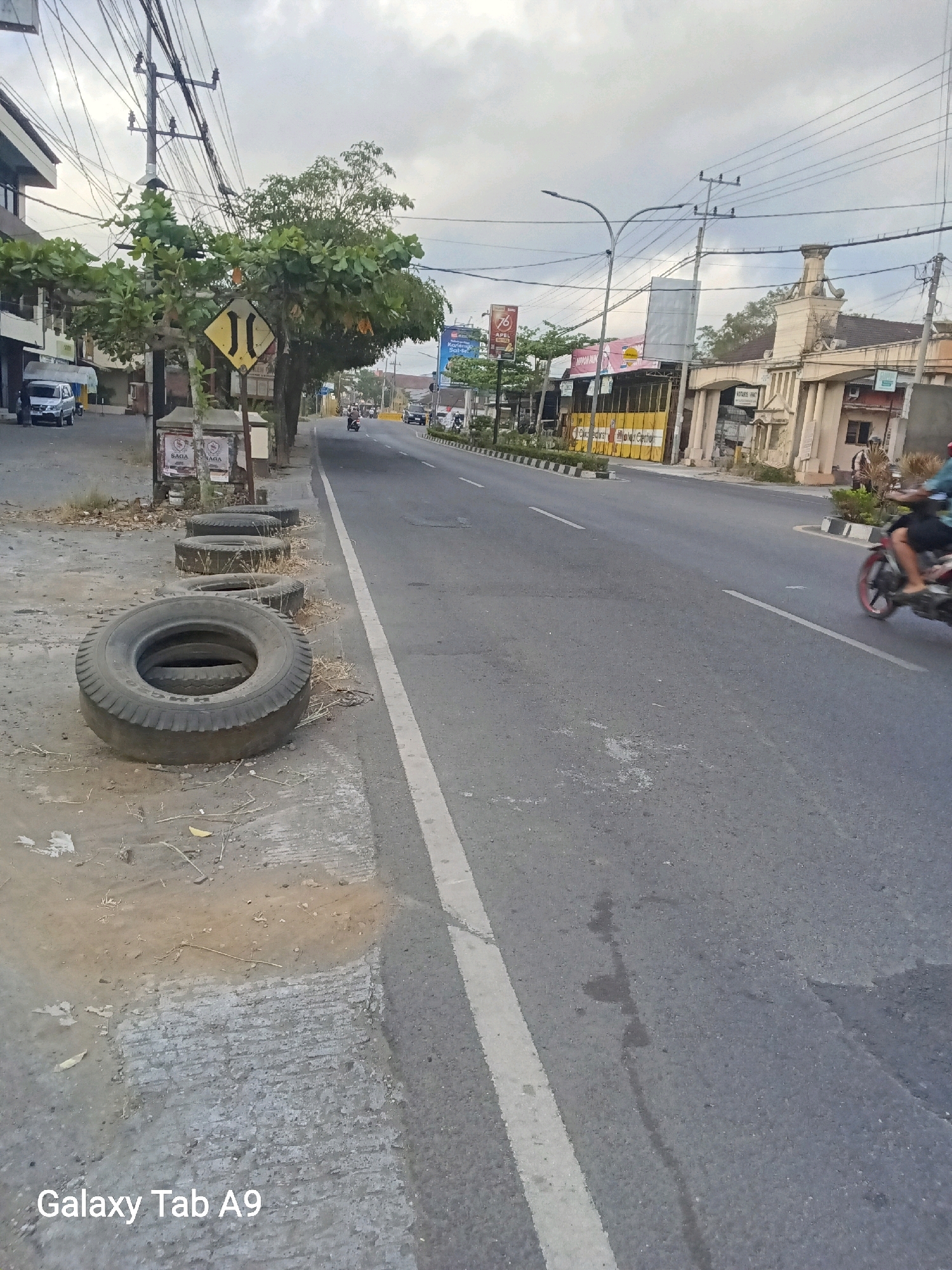 This image depicts a street scene in Trenggalek, Indonesia. On the left side of the image, a sidewalk is lined with several used tires, some stacked and one lying flat on the ground. Beyond the sidewalk, a building is visible with parked cars and trees. A yellow and black diamond-shaped traffic sign with two black vertical lines is mounted on a pole. The street itself is paved with asphalt and has a white lane marker. Several trees line the right side of the road. In the background, buildings and power lines are visible. A person is riding a motorcycle on the right side of the street. The sky is overcast. The camera's watermark, "Galaxy Tab A9," is present in the lower left corner of the image.
