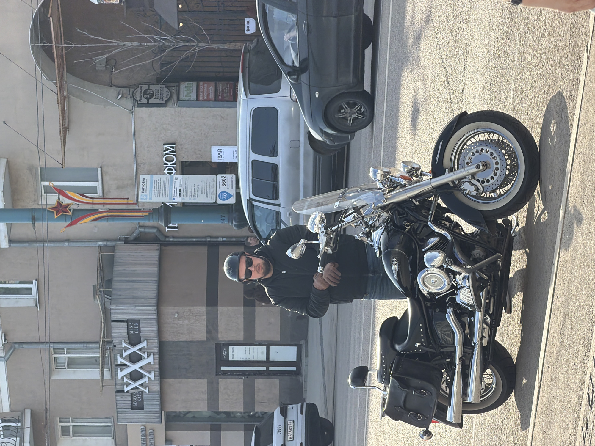 Here is a detailed description of the image:
**Scene:** The image shows a man standing next to a black motorcycle on a city street. He is wearing a black jacket, helmet, and sunglasses, and he appears to be looking at the motorcycle. The motorcycle is parked on the side of the road.
**Location:** The street is in Voronezh, Russia. Several buildings and signs indicate this location:
* A sign reads "ФЮМ" which is likely a local business name.
* Other signs are in Russian.
* A sign for "XXX MEN'S CLUB"
* A sign with the license plate "E414KX 36" which indicates a Russian license plate from the Voronezh region.
**Objects:**
* Motorcycle: A black motorcycle with saddlebags.
* Cars: Several cars and vans are parked nearby.
* Signs: Various signs are visible on the buildings, including business names and parking information.
* Street Decorations: A street light is decorated with a red star and yellow stripes.
* Building: Buildings with windows and doors are on the left and in the background.
**People:**
* Man: A man in a black jacket, helmet, and sunglasses.
* Possible other person behind the wheel of the vehicle.
**Actions/Interactions:** The man is standing next to the motorcycle, possibly admiring it or preparing to ride.
**Environment:**
* The scene is outdoors during the daytime.
* The road appears to be paved.
* The overall setting is urban, with buildings and vehicles indicating a city environment.