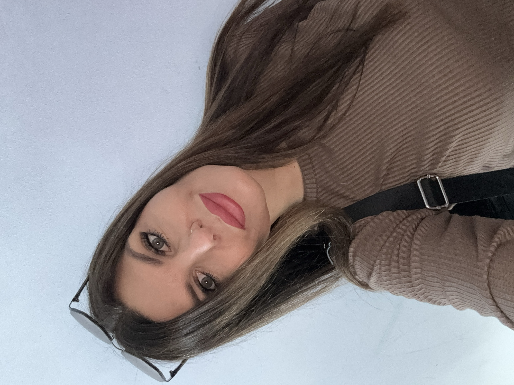 A woman is taking a selfie with a phone. She has long, dark brown hair, green eyes, and is wearing dark eyeliner and lipstick. She is wearing a brown, ribbed sweater and a black strap with a silver buckle is visible across her chest. Her sunglasses are resting on top of her head. The background is a light blue wall. There is no indication of the city of Chaikovsky, Russia in the image.