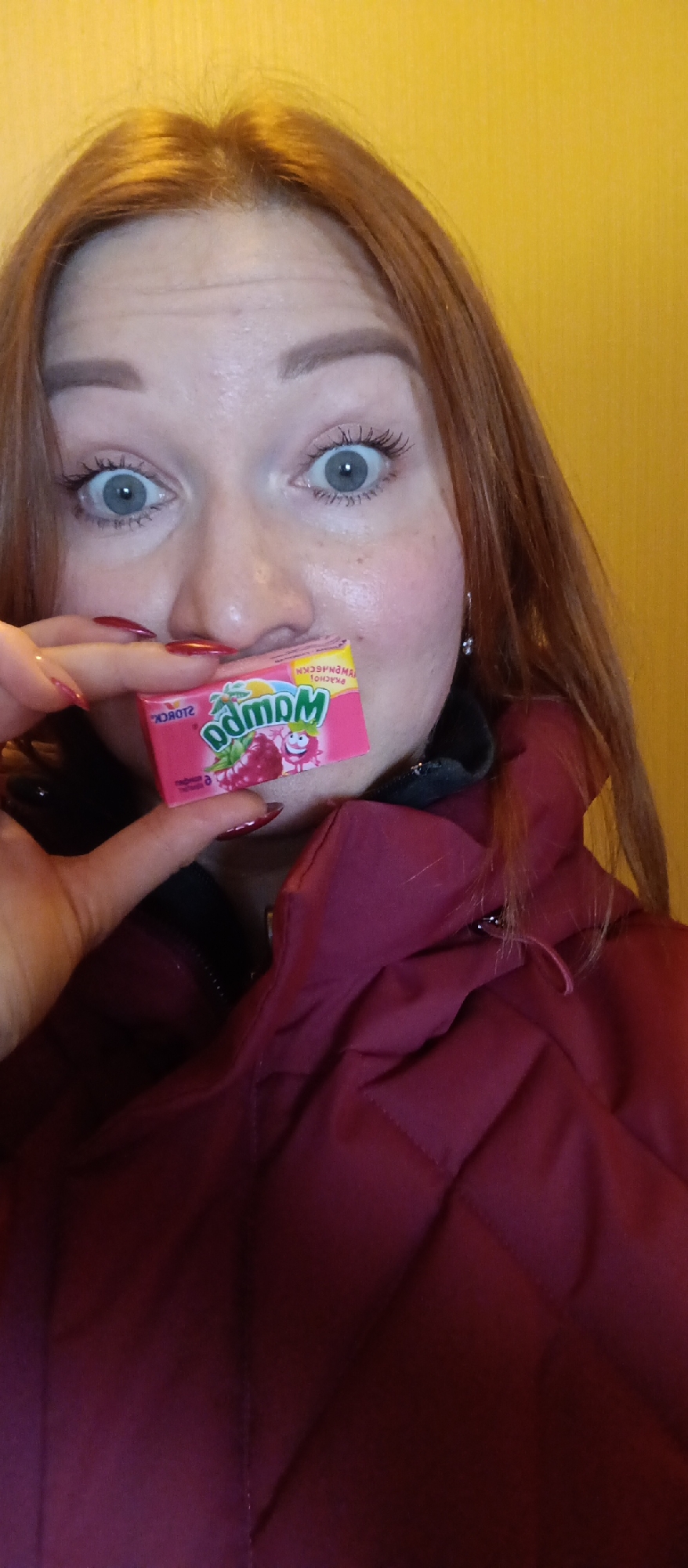 A close-up, portrait-oriented image shows a person, likely female, with reddish-brown hair and wide-open, light blue-grey eyes, conveying a surprised or playful expression. The person is wearing a burgundy or maroon puffy jacket. Their hand, with red painted fingernails, holds a small, pink rectangular package of candy or gum directly in front of their mouth.
The package prominently displays the brand name "MamBa" in stylized green font, along with the "STORCK" logo. Cyrillic text is visible, including "МАМБА," "МАЛИНА" (Raspberry), "ЖЕВАТЕЛЬНАЯ КОНФЕТА" (chewing candy), and "ТУТТИ" (Tutti Frutti). The packaging features illustrations of raspberries and a smiling cartoon fruit character. An earring is visible on the person's right ear. The background is a plain, light yellow wall. The lighting suggests an indoor setting, with no discernible time of day or specific location cues beyond an interior space.
Location context: Unknown, Unknown
