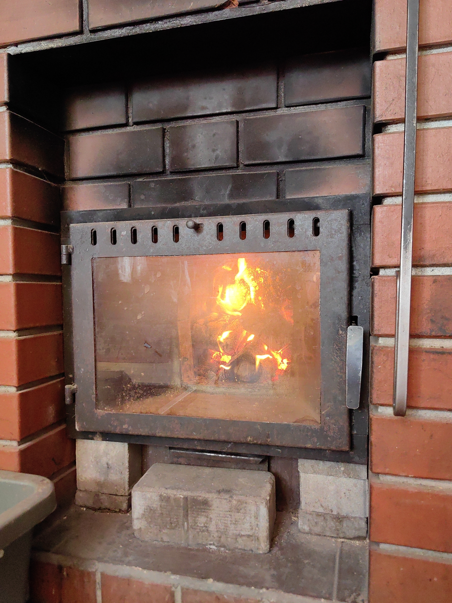 The image shows a close-up view of a fireplace within a brick structure, likely in a residential setting. The fireplace is made of dark metal with a glass door that allows a view of the burning fire inside. The fire is composed of glowing embers and flames, with visible logs. The surrounding area is constructed from red bricks, with some lighter colored bricks or concrete blocks used as supports beneath the fireplace. A metal handle is attached to the right side of the fireplace door, and a thin metal rod is positioned vertically along the right edge of the brick wall. The overall scene suggests a functional fireplace, possibly in use for heating or ambiance.