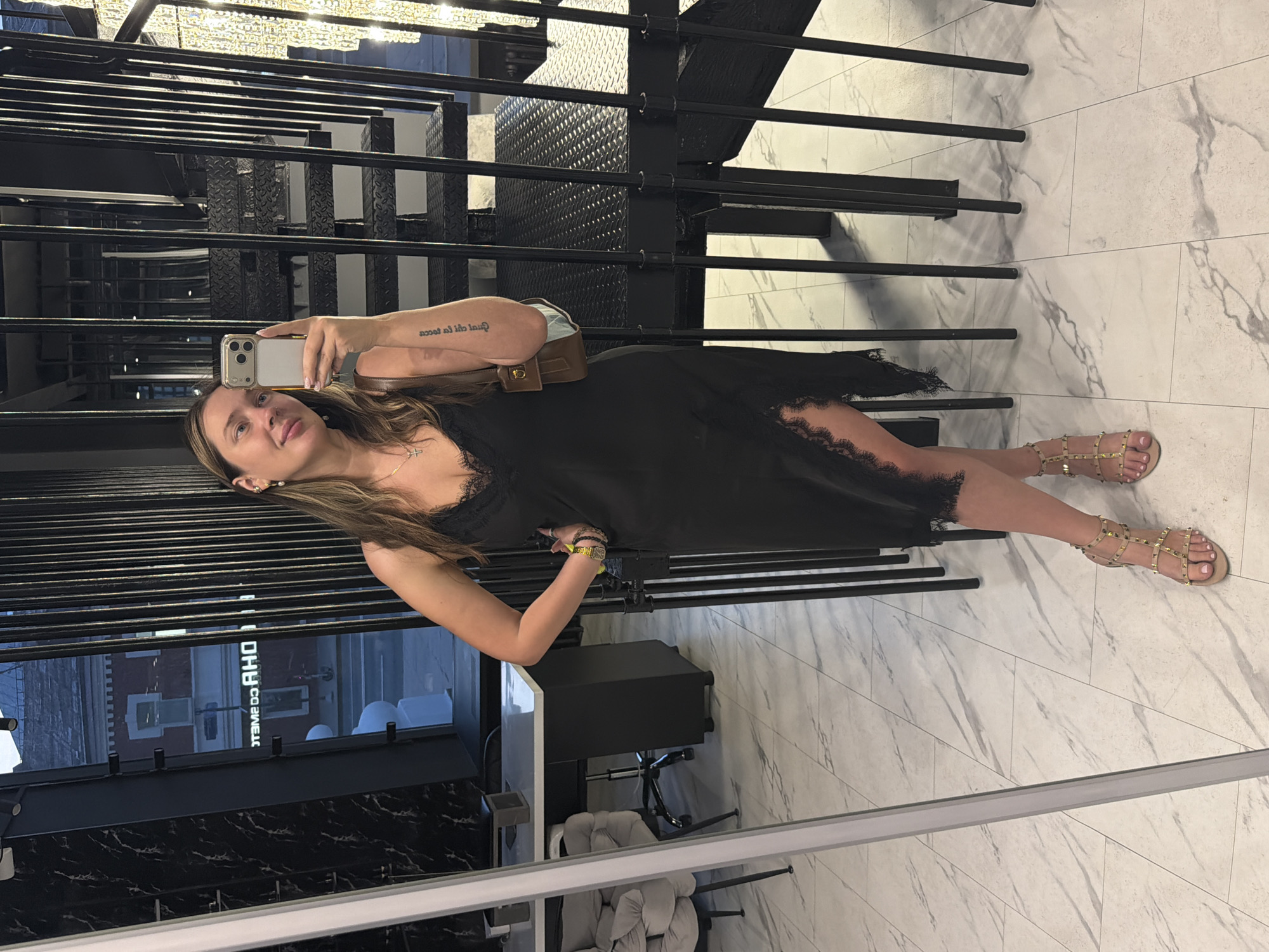 A woman is taking a mirror selfie in an indoor location with a modern, minimalist aesthetic. She is wearing a black slip dress with lace trim and a high slit, paired with gold studded sandals. She has a small brown crossbody bag and is wearing jewelry, including earrings and a necklace. In the background, there is a black metal staircase and a large chandelier with bright lights. The floor is tiled with a marble pattern. Through a window on the left, a street scene with buildings is visible.