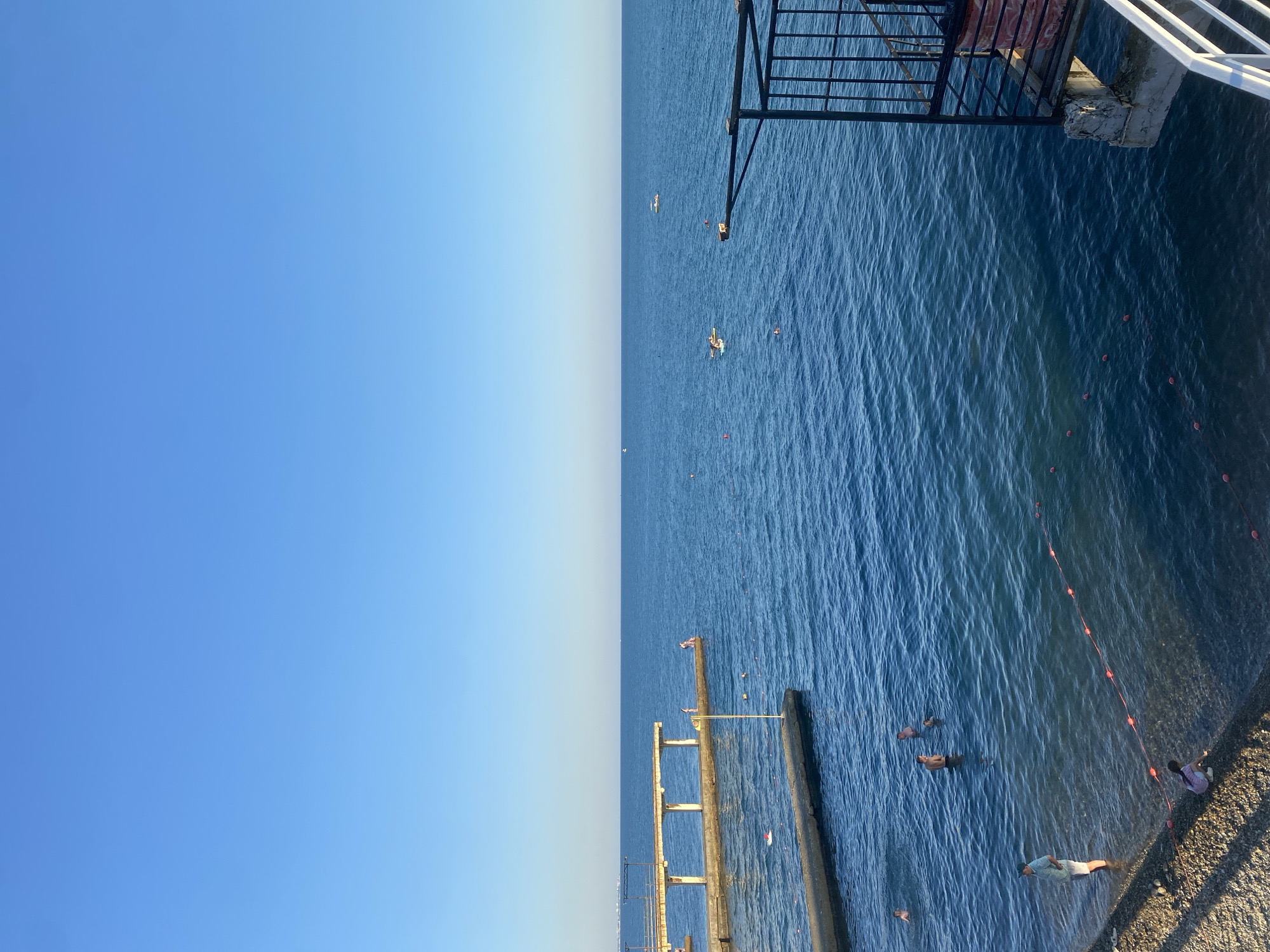 This media file depicts a serene scene at a beach in Sochi, Russia. The image is divided into two distinct halves: the left side is dominated by a clear, gradient blue sky, suggesting a sunny and pleasant day, possibly late afternoon due to the soft light. The right side showcases the calm, rippling blue water of the sea, meeting a pebbly shore.
Several people are visible enjoying the water. Some are swimming or wading near a concrete pier, while others are lying on the shore. A few individuals appear to be floating in the water, indicating a relaxed atmosphere. In the background, there are some small boats or rafts scattered on the water, and a dark structure, possibly a lifeguard tower or a viewing platform, is visible at the top right. A white railing, part of a structure extending over the water, is also present in the upper right corner. Orange buoys create a boundary line in the water, marking a designated swimming area. The overall mood is peaceful and recreational. There is no visible text in the image.
