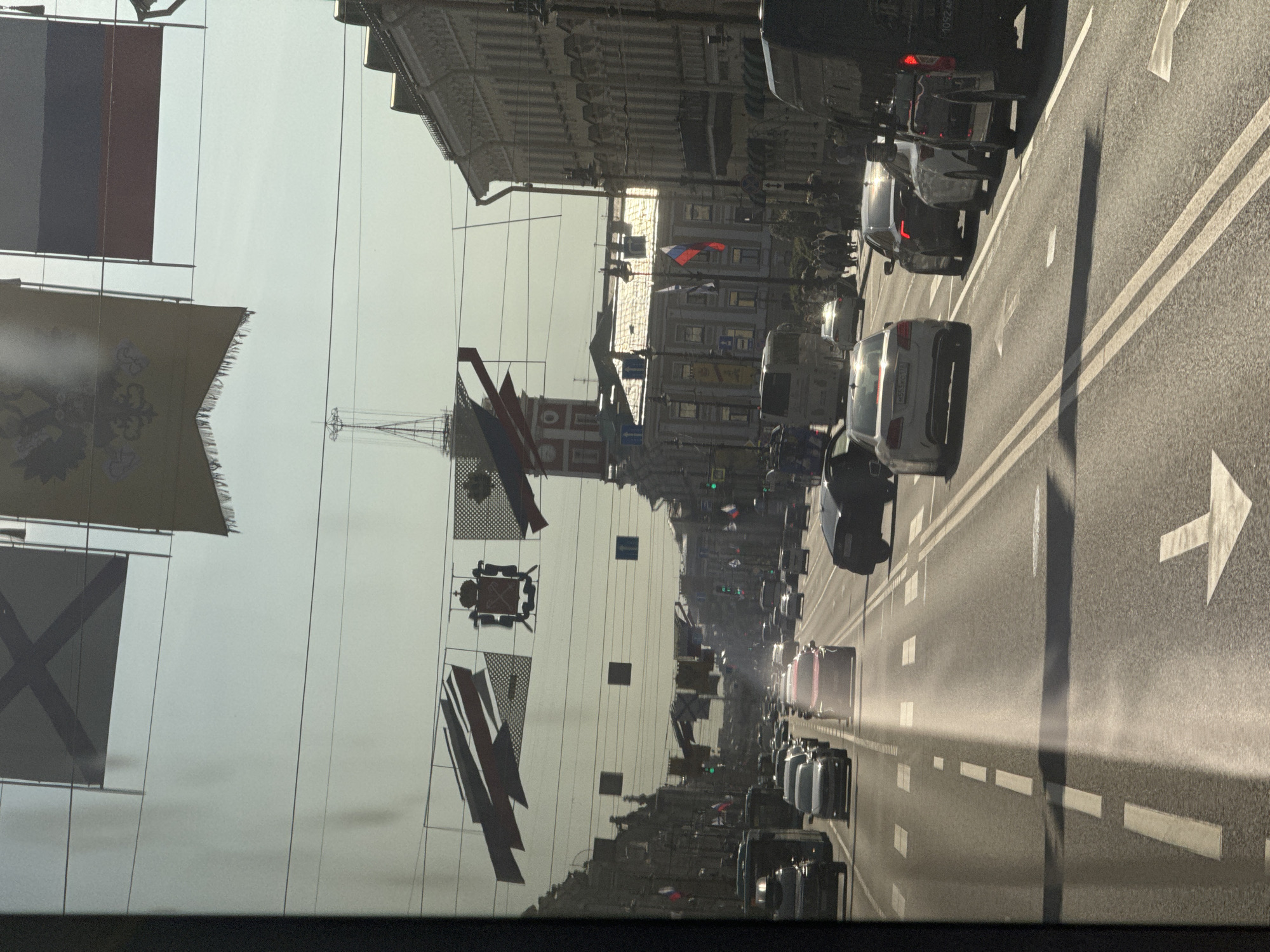 The image shows a view from a vehicle looking down Nevsky Prospekt in Saint Petersburg, Russia. The street is lined with classical architecture and decorated with numerous flags and banners, indicating a festive occasion or parade. The flags are a mix of national and historical designs, including the Russian tricolor and emblems with double-headed eagles.
Traffic is moderate, with cars and a bus on the multi-lane road. Pedestrians are visible on the sidewalks. The sun is shining, casting long shadows across the street, suggesting it is either morning or late afternoon. The overall atmosphere appears to be one of a bustling urban environment during a public event.