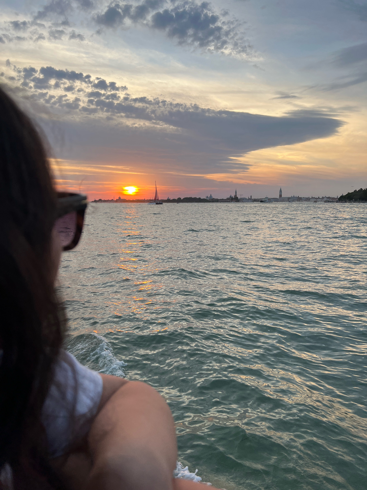 A person, seen from behind, with dark hair and wearing sunglasses and a white shirt, is looking out at the water. The scene is of the Venice lagoon at sunset. The sun is setting on the horizon, casting a warm glow on the sky and the water. A sailboat is visible in the distance, along with the silhouette of the Venice skyline, including buildings and church towers. The water is rippled, reflecting the colors of the sunset. The image appears to be taken from a moving boat, as indicated by the wake at the bottom left.