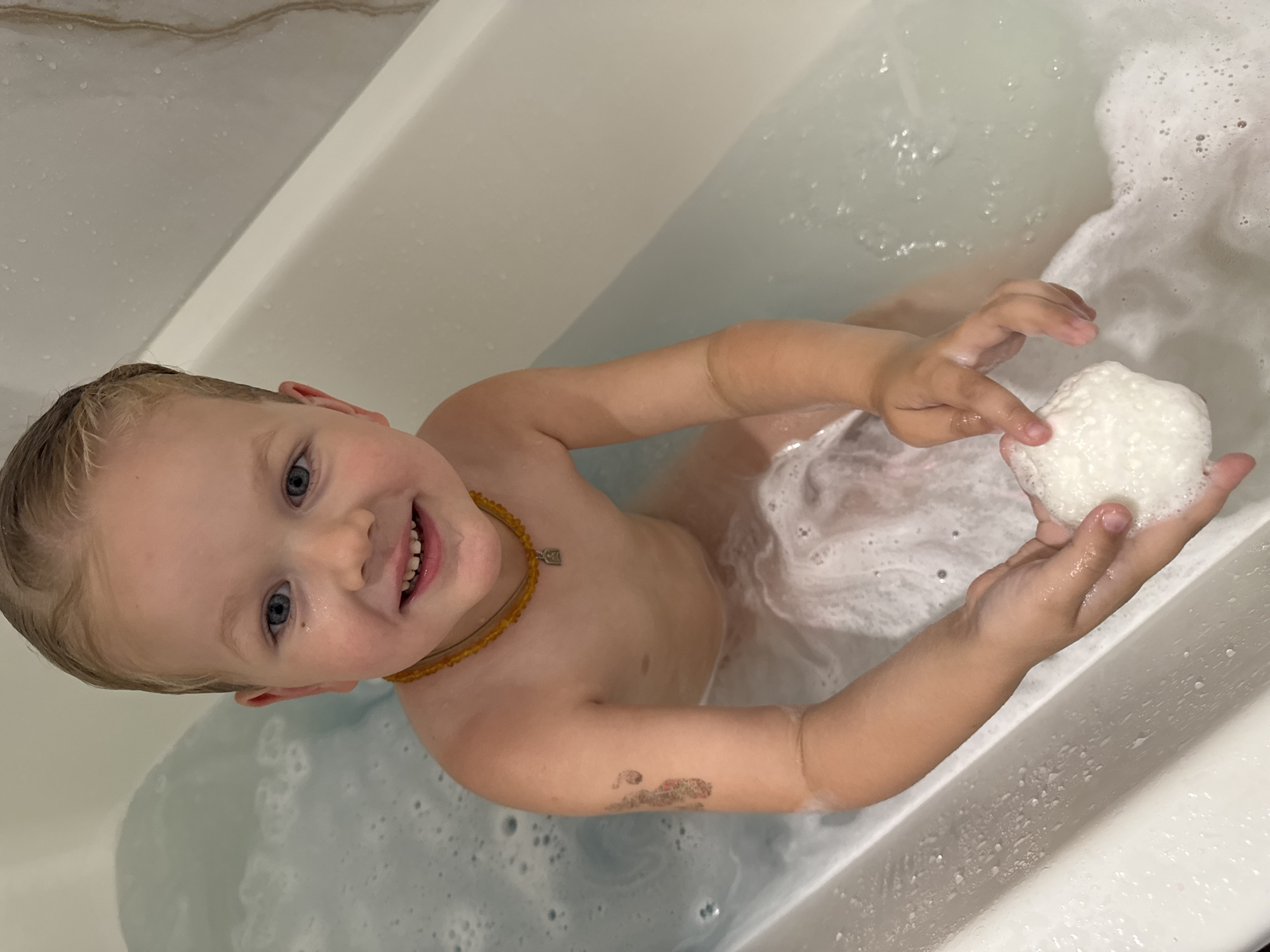A young boy with blonde hair and blue eyes is sitting in a bathtub filled with bubbly water. He is wearing an amber necklace. His left arm has a birthmark or tattoo of a cartoonish footprint. He is holding a white, textured object, possibly a bath bomb or a toy, in his hands. The bathtub is white and appears to be made of porcelain or acrylic. The background shows a wall with subtle veining, suggesting it is tiled or made of a similar material. The lighting is soft, indicating an indoor setting. The scene is focused on the boy and his interaction with the bath products, suggesting a moment of play or relaxation during bath time. The location is described as Ферма, Russia, but this information cannot be verified from the image itself.