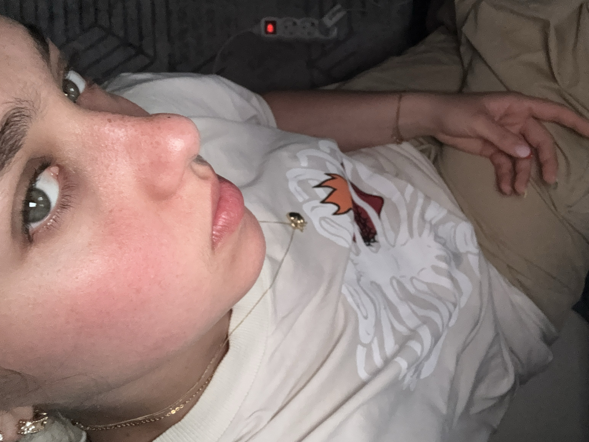 A selfie-style close-up photograph shows a person lying on a bed or couch with a patterned shirt. The person has fair skin, green eyes, and light-colored hair. They wear a gold necklace and a gold bracelet. The shirt features a design with white and beige abstract floral patterns. A cable and a power strip are visible in the background. The setting appears to be indoors. The photo does not provide information about the location in Milan, Italy.