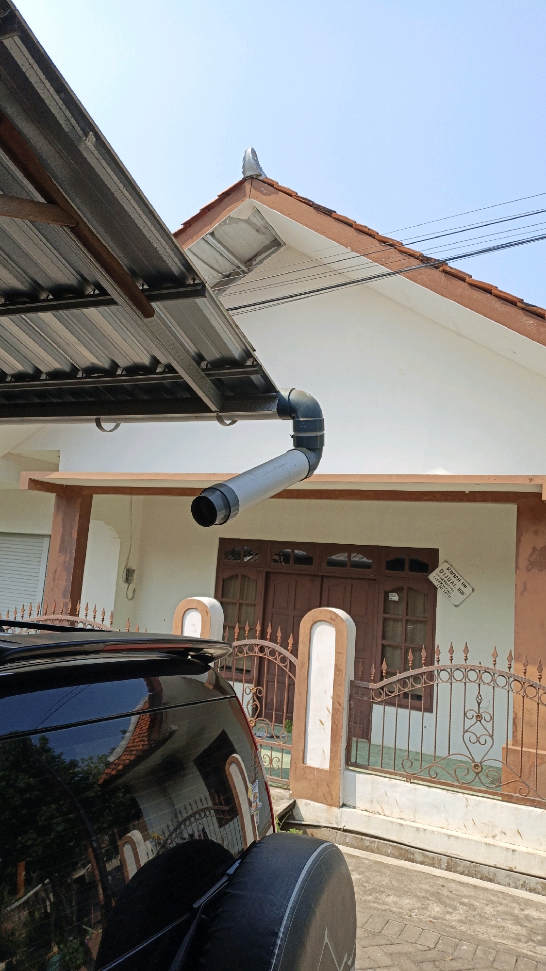 A black SUV is parked in front of a house with a brown roof and white walls in Cikal, Indonesia. A downspout extends from the roof over the driveway. A metal fence with decorative scrollwork separates the property from the street. The sky is clear and blue.