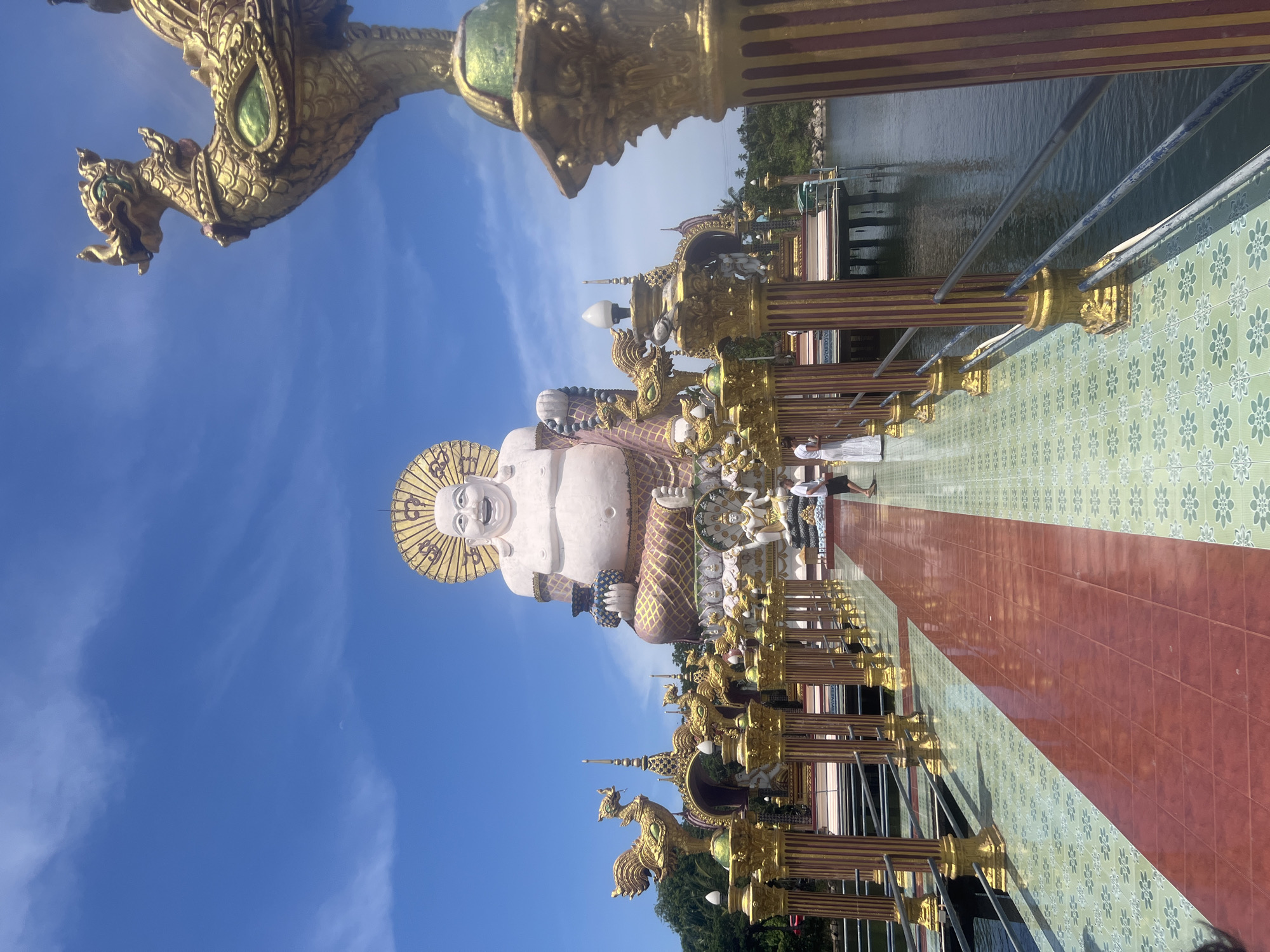 A wide, low-angle shot captures the Big Buddha of Ko Samui, Thailand, on a sunny day with a clear blue sky. The colossal white statue of a seated Buddha with a halo dominates the background. In front of it, a pathway made of red and green tiles leads towards the statue, bordered by ornate golden pillars topped with bird-like sculptures. Two individuals, one in white and one in black, are walking on the pathway, adding a sense of scale to the immense Buddha. The pathway extends over calm, dark water, with lush green trees and distant land visible in the background. The overall scene is one of grandeur and religious significance.