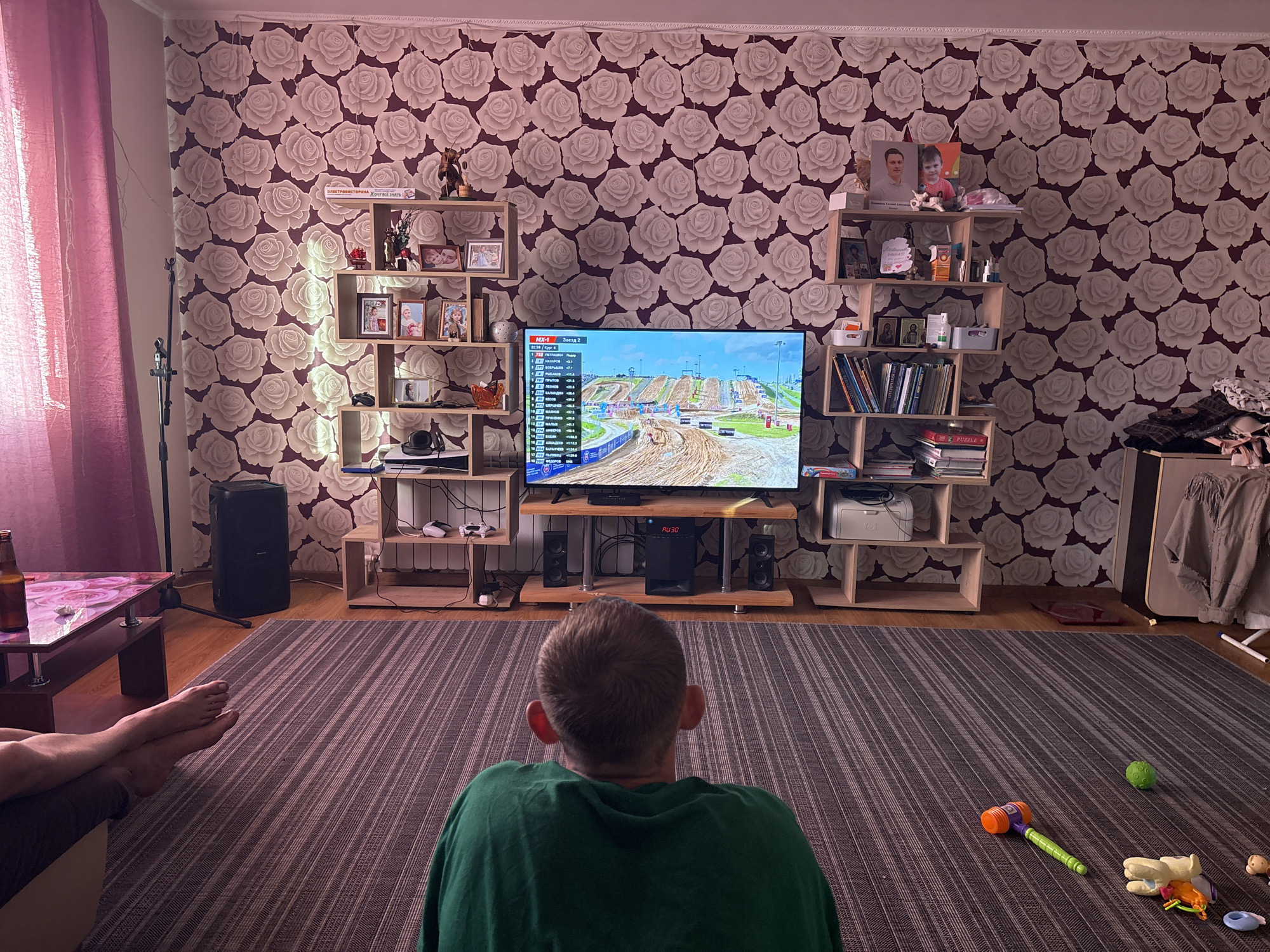 The image captures an indoor domestic scene, likely a living room, in Votkinsk, Russia. The central focus is a man, viewed from behind, wearing a green t-shirt, seated on a dark striped rug and intently watching a large flat-screen television. The TV displays a motocross race, featuring a digital scoreboard on the left with Cyrillic text indicating "MX-1" and "Заезд 2" (Race 2), along with rider names and statistics.
To the left of the frame, a person's bare feet, with painted toenails, are visible, crossed at the ankles and resting on an unseen surface, suggesting relaxation on a sofa or chair. Adjacent to these feet is a small, dark wooden table with a glass top that exhibits a pink floral design underneath; a brown glass beer bottle rests on it. A tall, black floor-standing speaker and a black microphone stand are positioned in the left corner of the room.
The background wall is covered with wallpaper featuring a repeating pattern of large, light-colored roses on a dark maroon base. Flanking the television are two asymmetrical, light wooden shelving units. The left unit contains various framed photographs (some featuring children), books, and small decorative items. The right unit holds additional framed photographs (including a prominent image of a man and a child), books (one with a visible Cyrillic title: "ЭЛЕКТРОННАЯ ТОРГОВЛЯ. ХОЧУ ВСЁ ЗНАТЬ," meaning "Electronic Commerce. I WANT TO KNOW EVERYTHING."), religious icons, and other decorative objects.
Below the television, on a matching wooden stand, a black central unit with the text "AU30" is visible, flanked by two smaller black speakers. Gaming controllers (white) are placed on a lower shelf of the TV stand to the left, and a white printer is on a lower shelf to the right. In the far right of the room, clothing items, including a brown jacket, are draped over a light-colored wooden storage unit. Scattered on the dark striped rug in the foreground are several children's toys, including a green textured ball, an orange and purple toy hammer with a green handle, a white animal-shaped toy, a small doll, and a plastic ring. The floor visible around the rug's edges is made of wooden laminate or parquet.