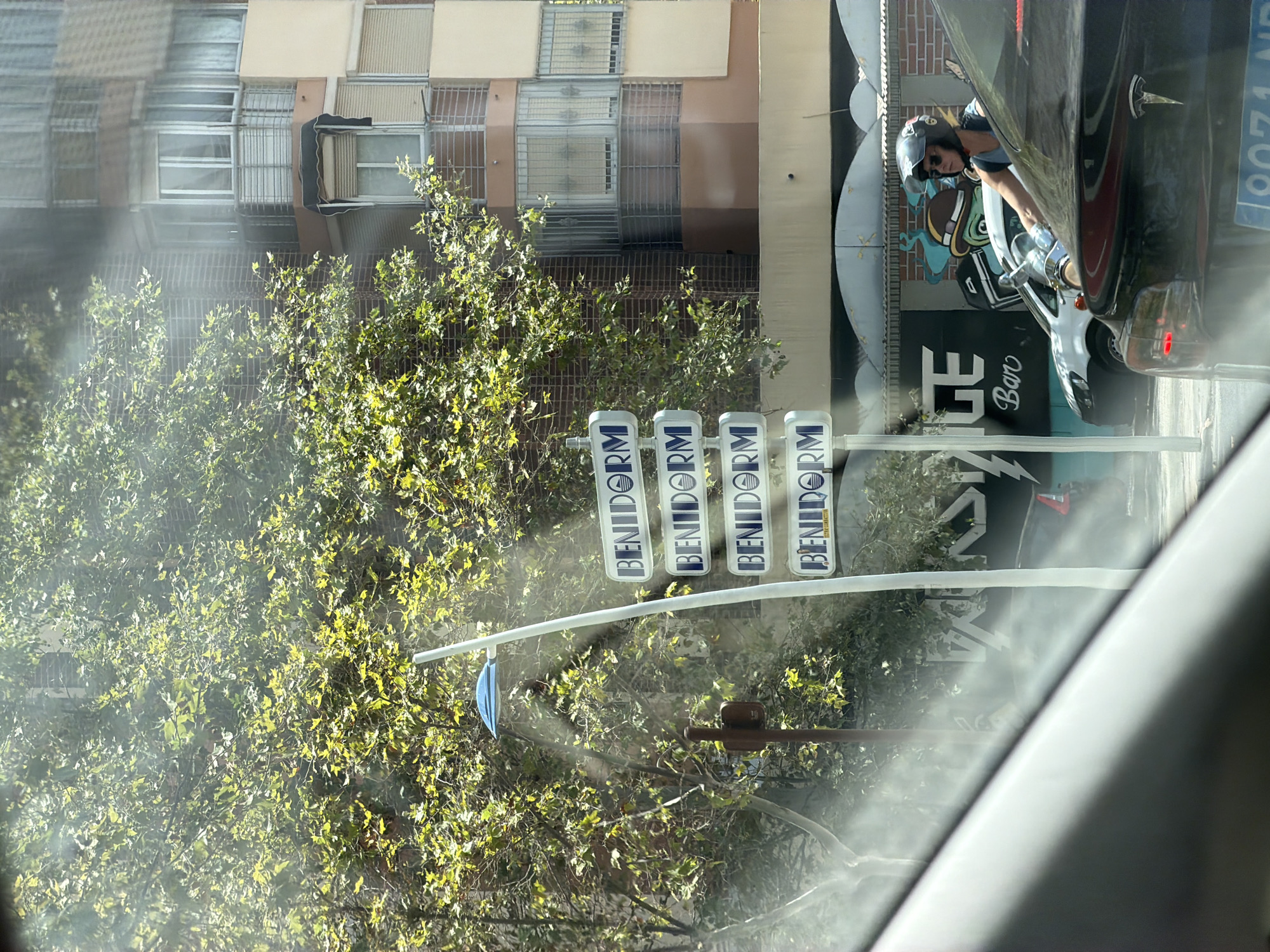 The image depicts a street scene in Benidorm, Spain, characterized by a clear, bright sky and lush green foliage. A modern, multi-story building with balconies and windows is visible in the background. In the foreground, a street lamp with a blue light fixture stands near a directional sign that repeatedly displays "BENIDORM". The sign appears to be mounted on a pole. Further down the street, a woman wearing a helmet and sunglasses is riding a motorcycle, moving through traffic. She is partially obscured by a black car in the immediate foreground, the rear of which shows a Tesla logo and a license plate. Beside the motorcycle, a white car is also visible. In the background behind the motorcycle rider, there is a building facade with graffiti-style artwork that includes the word "Bar". The overall impression is that of a sunny day in a Spanish city, likely a tourist destination given the prominent signage.