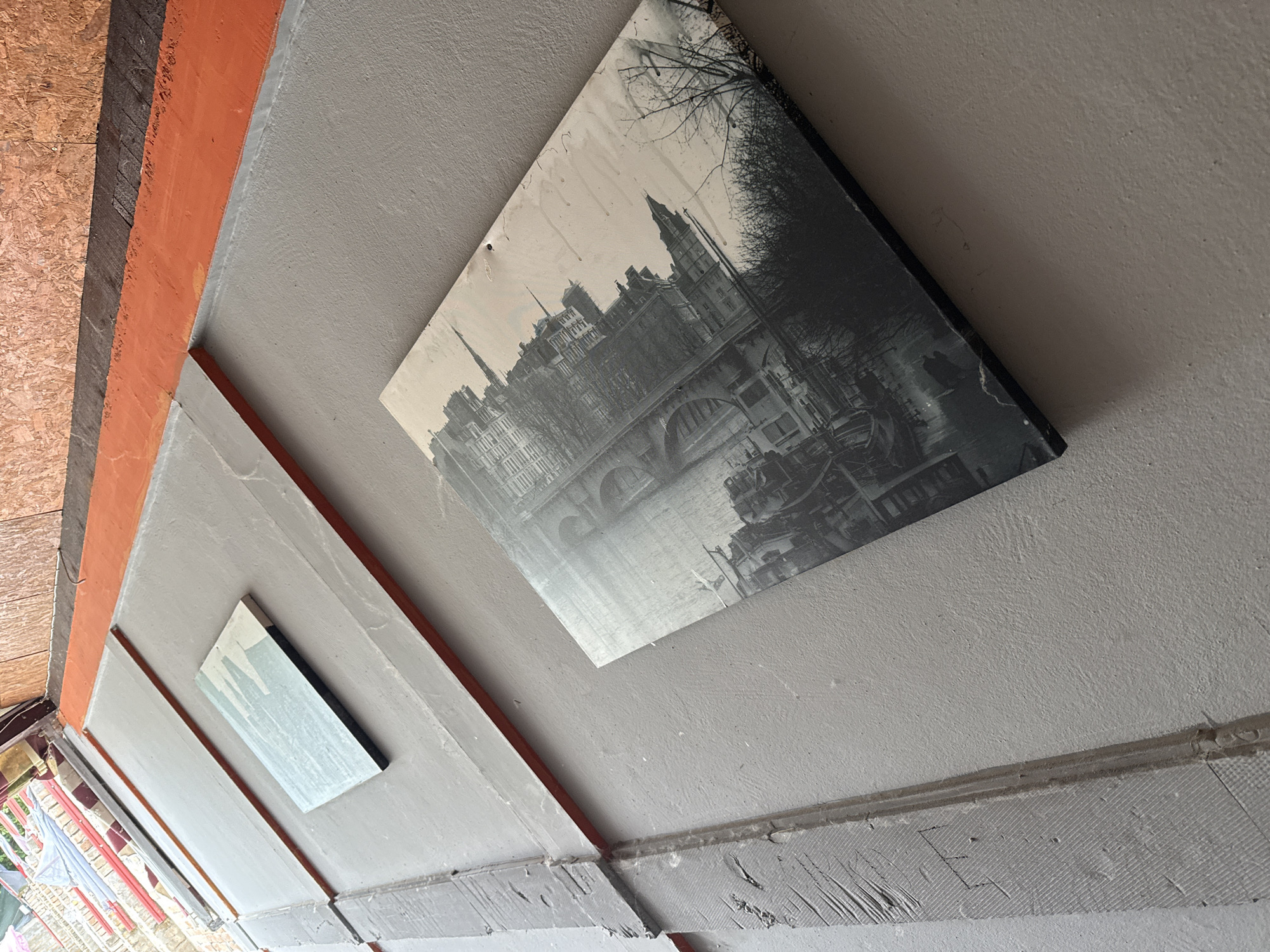 The image displays a section of an exterior wall, likely part of a building's facade. The wall is painted a neutral gray and features several rectangular elements with orange trim. Two of these elements are visible: one smaller, lighter-colored rectangle, and a larger, diamond-shaped black and white photograph of a cityscape. The photograph appears to be of an older European city with many spires and intricate architecture, possibly an industrial or historic district, with bare trees in the foreground. The wall itself has a textured finish and shows signs of weathering and wear, including some unevenness in the paint and grout lines. The scene suggests an outdoor urban environment. Based on the provided context, the location is identified as the city of Apatin Municipality, Serbia.