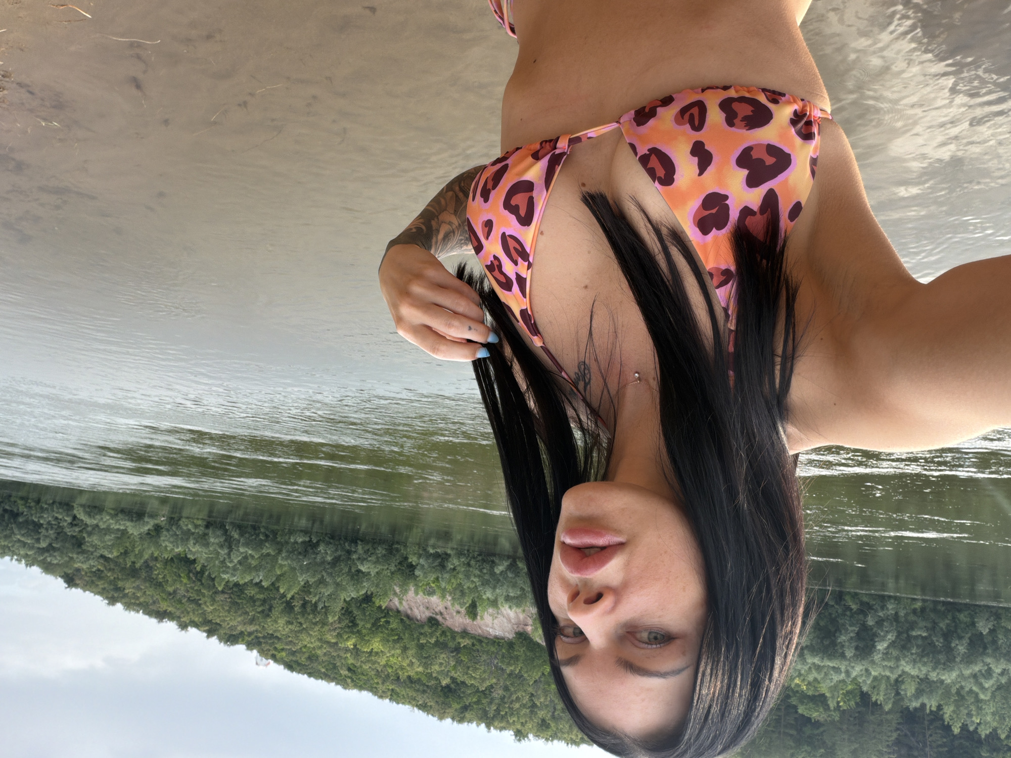 A woman with dark hair and a leopard-print bikini stands on the bank of a wide, calm river. She is looking towards the camera and has her hand near her hair. The riverbank is sandy and the water is shallow near the edge. In the background, a lush green forested hillside rises steeply from the opposite bank of the river. The sky is overcast. The scene appears to be located along the Ufa River in Ufa, Russia.