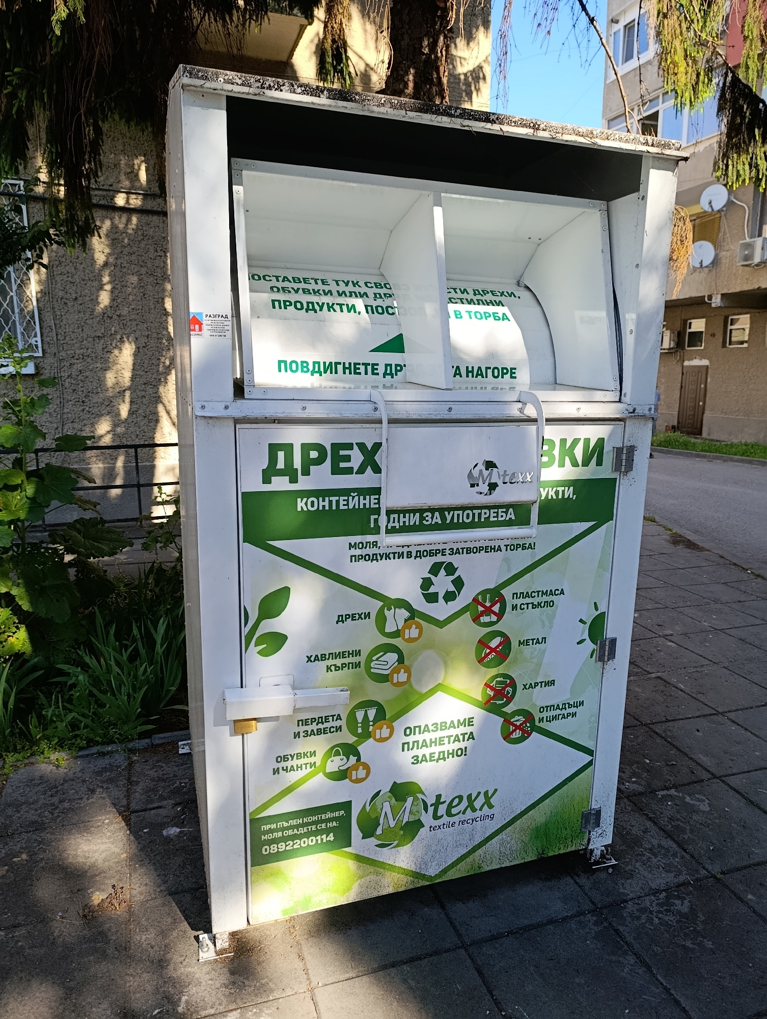 The image shows a white textile recycling bin located in front of a building with a tree to the left and a building with windows and satellite dishes to the right. The bin has green lettering and graphics. The top section of the bin has an opening with a white interior, and the sides of the opening have text. The main body of the bin has text and icons indicating what can be recycled, such as clothes, shoes, bags, plastics, metal, and paper. There is a logo for "Mtexx textile recycling" on the bin. There is also a label with the text "РАЗГРАД" which indicates the city of Razgrad, Bulgaria. The bin is situated on a paved surface. The lighting suggests it is daytime.