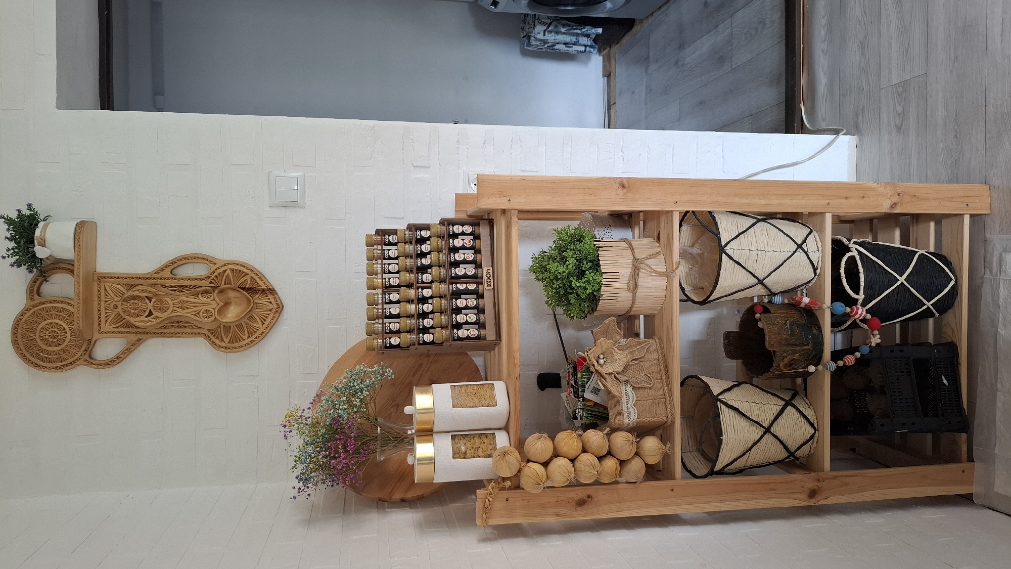 This is a full-shot, eye-level, indoor photograph of a wooden shelving unit filled with various decorative and practical items. The shelving unit is placed in a corner with a white brick-patterned wall behind it.
On the top shelf, a wooden wall decor piece with intricate carvings is mounted, featuring a heart shape at the bottom. A small white pot with a sprig of lavender sits on a small shelf attached to the decor. Below this, on the top shelf of the unit, are two tall, white jars with gold lids containing pasta, and a tiered wooden spice rack filled with numerous small spice jars. To the left of the jars, a vase of dried, multi-colored baby's breath flowers is visible.
The second shelf down holds a string of garlic bulbs hanging on the left. In the center, there's a small woven basket with a burlap flower decoration and another decorative element with a tag. To the right, a potted green plant in a wooden cylinder-like container is present, tied with twine.
The third shelf from the top contains two woven baskets with black criss-cross patterns. Between them sits a dark, rustic-looking pot with a colorful beaded garland draped around it.
The bottom shelf has a black plastic crate filled with items, possibly potatoes or other produce. To the right of the crate, another woven basket with a similar black criss-cross pattern is present, this one being black with white accents.
The floor appears to be light gray laminate flooring. To the right of the shelving unit, a doorway leads to another room, where a washing machine and some laundry are visible. The overall impression is one of a cozy and organized kitchen or pantry area.
The image is described as being from Cherkessk, Russia. However, there are no visual elements in the image that specifically identify it as being in Cherkessk, Russia. The description is based solely on the visual content of the image.