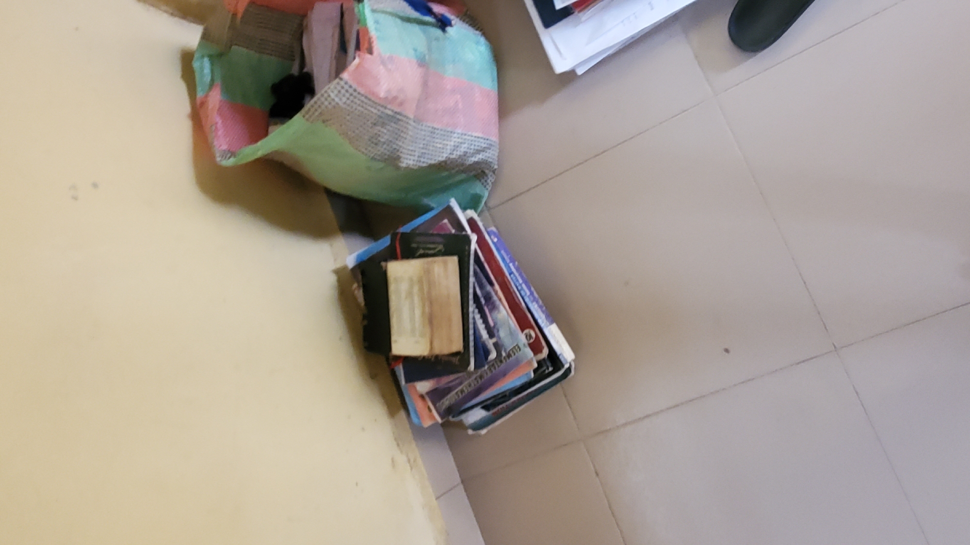 In Jalingo, Nigeria, a pile of books rests on a beige tiled floor next to a wall. The books are stacked haphazardly, with some open and others closed. A large, colorful bag, possibly a tote bag, is situated to the right of the books. The bag appears to be made of fabric with a patchwork design featuring green, pink, and white squares. It is partially filled, with some black material visible inside. In the background, a light yellow wall extends upwards, showing some minor imperfections. The overall scene suggests a casual, perhaps domestic or academic, setting.