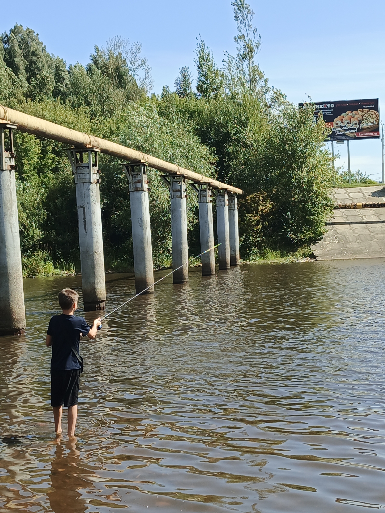 A young person, viewed from behind, stands in shallow water and fishes. The person is wearing a dark blue t-shirt and black shorts. They are holding a fishing rod with the line cast into the water. The water appears to be a river or canal, with ripples on its surface. In the background, a large pipe supported by concrete pillars runs horizontally across the scene. Beyond the pipe and pillars, lush green trees and bushes form a dense treeline. A billboard advertising a food establishment is visible on the right side of the image, behind the trees. The sky above is clear and blue. The scene appears to be located near an industrial area, possibly on the outskirts of Zlatoust, Russia, given the presence of industrial infrastructure and the specific billboard design.