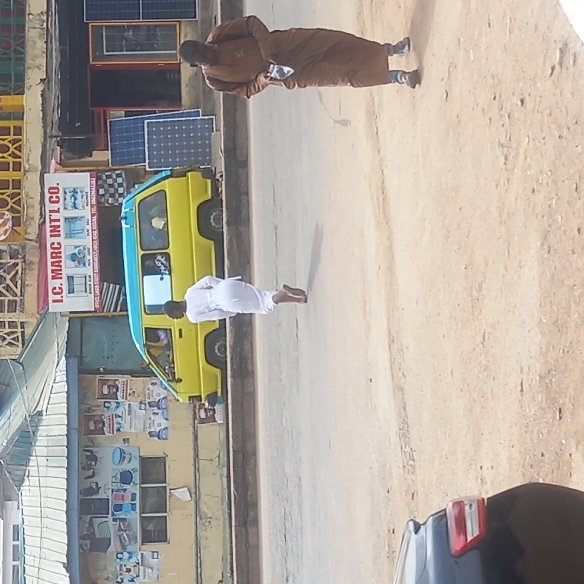 This outdoor street scene, likely set in Sardauna, Nigeria, captures a moment on a bright, sunny day. The image is oriented vertically but depicts a street running horizontally across the frame.
The central subjects are two individuals and a yellow van. On the right side of a paved road, a person dressed in a long, brown traditional robe stands facing left, holding a light-colored object, possibly a phone, in their hands. Further down the road to the left, another person, clad in a white traditional robe, walks away from the viewer.
A distinctive yellow van with a light blue top is parked alongside the road on the left. The vehicle's windows reflect parts of the surroundings. To the far left, a building serves as a backdrop, featuring a sign for "I.C. MARC INT'L CO." Visible below this are the words "BAND BALI" and a partial address and phone number: "OFFICE: LAGES STREET ROUNDABOUT, ABUJA AGAD KUDUSAL TEL". The storefront displays several solar panels, both mounted on the building and resting on the ground, suggesting the nature of the business. Various posters are also affixed to the building's wall.
The ground consists of a paved road and a drier, sandy shoulder on the right. The bright daylight creates clear shadows, indicating midday conditions. In the bottom right corner, the rear section of a dark-colored vehicle, showing a red taillight, is partially visible. The overall scene suggests a commercial area with daily activity.