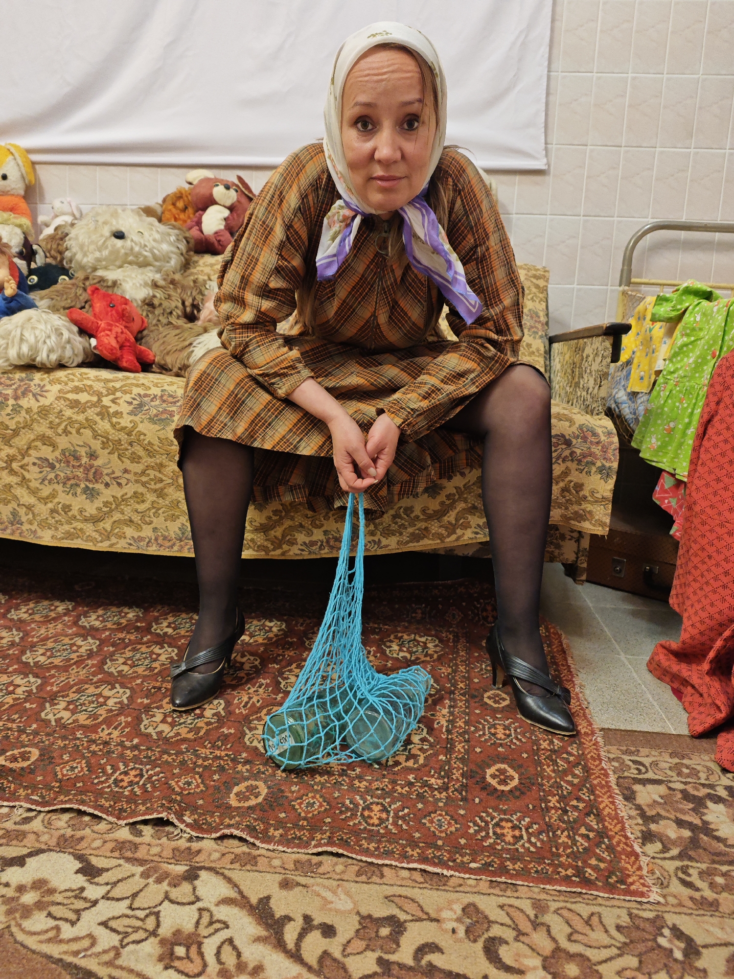 A medium shot captures a woman sitting on the edge of an old-fashioned couch in a cluttered indoor setting. She has fair skin, light brown hair partially visible under a cream-colored headscarf with purple and blue floral patterns, and is looking directly at the viewer with a serious, slightly questioning expression. She wears a long-sleeved plaid dress in brown, orange, and beige tones, dark sheer tights, and black high-heeled shoes with a strap design. Her hands are clasped around a blue mesh string bag (an "avoska") containing several clear glass jars or bottles; faint text, possibly "0.5L", is visible on one bottle.
The couch she sits on features a gold and brown floral patterned upholstery. Behind her, the couch is piled high with numerous stuffed animals, including a large shaggy brown bear, a bright red plush toy, and various others in different colors and textures. To her right, a metal-framed bed or cot is visible, draped with colorful, patterned clothes in green, yellow, and red. Below this, a dark brown suitcase or trunk rests on the floor.
The floor is covered by two layered rugs: an upper, larger red rug with intricate traditional patterns, and a lower, lighter beige/brown patterned rug, with the red rug's edge showing some fraying. The background wall behind the couch is partially covered by a white sheet, while the wall to the right features light-colored square tiles. The scene evokes a retro, possibly Eastern European or Soviet-era domestic atmosphere.