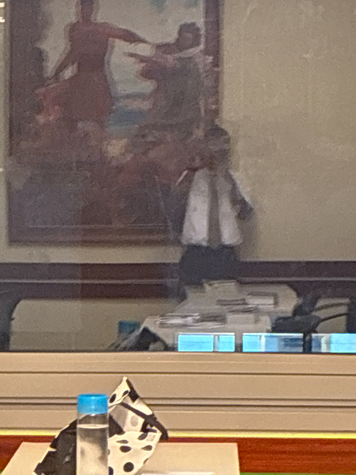 The image shows a reflection on a glass surface. In the background, a painting depicts two figures in a dance. In the foreground of the reflection, a person wearing a white shirt and tie stands behind a table with papers. Below the reflection, a bottle of water with a blue cap and a black and white polka dot mask are visible on a surface. The scene appears to be indoors, possibly in a public building or a school, based on the presence of a painting and educational materials. The location is identified as Accra, Ghana.