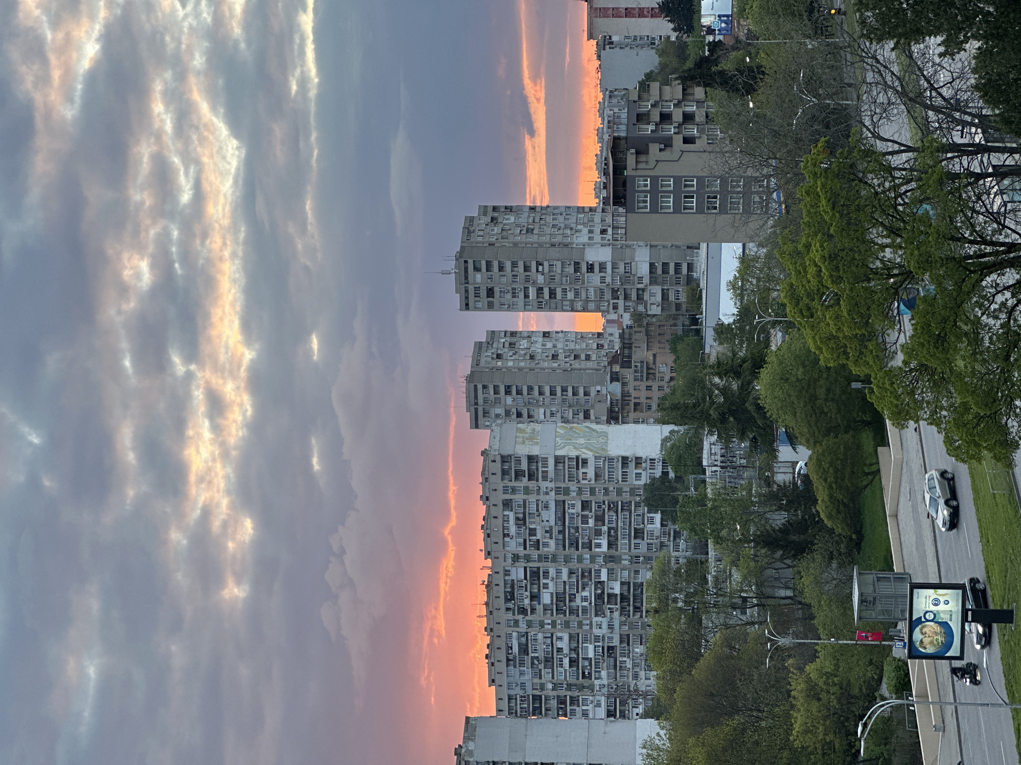 An urban landscape is captured during what appears to be either sunrise or sunset, distinguished by a dramatically colored sky. The image features a row of tall, multi-story residential buildings, likely apartment blocks, constructed from light grey concrete with numerous windows and balconies, many equipped with visible air conditioning units. One building in the midground displays a large, light-colored mural or abstract design on its facade.
The sky occupies the upper half of the frame, transitioning from soft grey-blue clouds with golden-peach highlights at the top to a vivid, intense band of orange and pink light along the horizon, indicating the low position of the sun. Abundant green trees with dense foliage are prominent in the foreground and midground, bordering a multi-lane asphalt road.
On the road, visible in the lower right, a light-colored sedan, possibly silver or white, is driving, and another darker vehicle is partially seen. A large street billboard stands prominently on the right side of the road. The billboard features an advertisement with a close-up image of a person's face and circular graphics. Visible text includes "Conmedica" and "Medicina," suggesting a medical or health-related service. Streetlights are also present. The scene depicts typical urban activity against a backdrop of a colorful sky, creating a striking contrast between the natural phenomenon and the built environment.