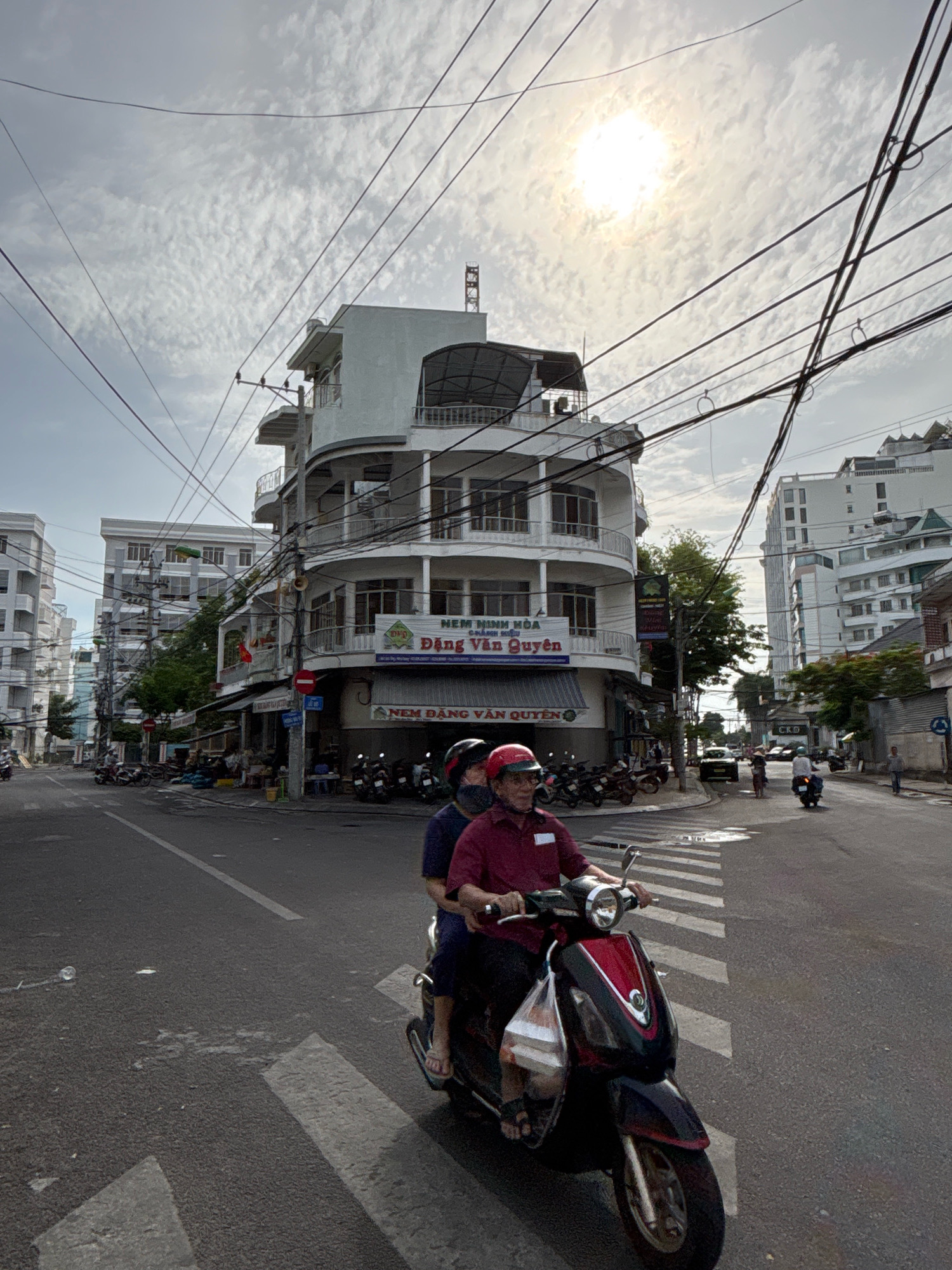 The image captures a street scene in Nha Trang, Vietnam, likely during daytime under a partly cloudy sky with the sun visible. The foreground features a red and black scooter with two individuals on it, one driving and one as a passenger. The driver wears a red collared shirt and a red helmet, while the passenger wears a dark blue shirt and a black helmet. They are riding on a paved road with a white crosswalk marking in the foreground.
Behind the scooter, a distinct multi-story building with a curved facade stands prominently. This building has multiple balconies and windows. Signs are visible on the building, one with Vietnamese text that includes "NEM MINH HOA" and another below it with "DONG VAN QUYEN". Adjacent to this building, there are other buildings and shops lining the street. Various motorbikes are parked along the sidewalk. Overhead, a complex network of electrical wires crisscrosses the sky.
Further down the street, other buildings of varying heights are visible, including modern high-rise structures. Traffic appears to be light, with a few other scooters and a car seen in the distance. The overall atmosphere suggests an urban environment in Vietnam. The specific location of the building with the curved facade and the signs could help pinpoint the exact street or intersection.