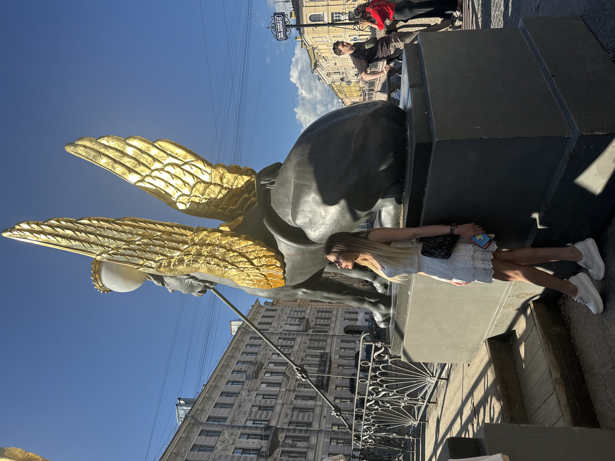 This media file captures a sunny day in Saint Petersburg, Russia, with a young woman posing next to a large, dark-colored statue adorned with golden wings. The statue appears to be a mythical creature, possibly a griffin or a winged horse, holding a staff with a glowing orb. The woman is wearing a white lace dress and white sneakers, and she is leaning against the statue's base. She has blonde hair and is wearing glasses. Behind the statue and the woman, a multi-story building with a classical architectural style is visible, featuring numerous windows and decorative elements.
In the background, other people are present, suggesting a public space, possibly a bridge or a square. One individual in a red top is partially visible, and another person in a dark shirt is leaning against a structure. The sky is a clear, bright blue, indicating fair weather and likely midday. A street sign with Cyrillic lettering is also visible, providing a clue to the location. The overall atmosphere is cheerful and relaxed, with the bright sunlight casting shadows and highlighting the golden details of the statue. The scene appears to be set outdoors, on a paved surface with some decorative metal fencing present.