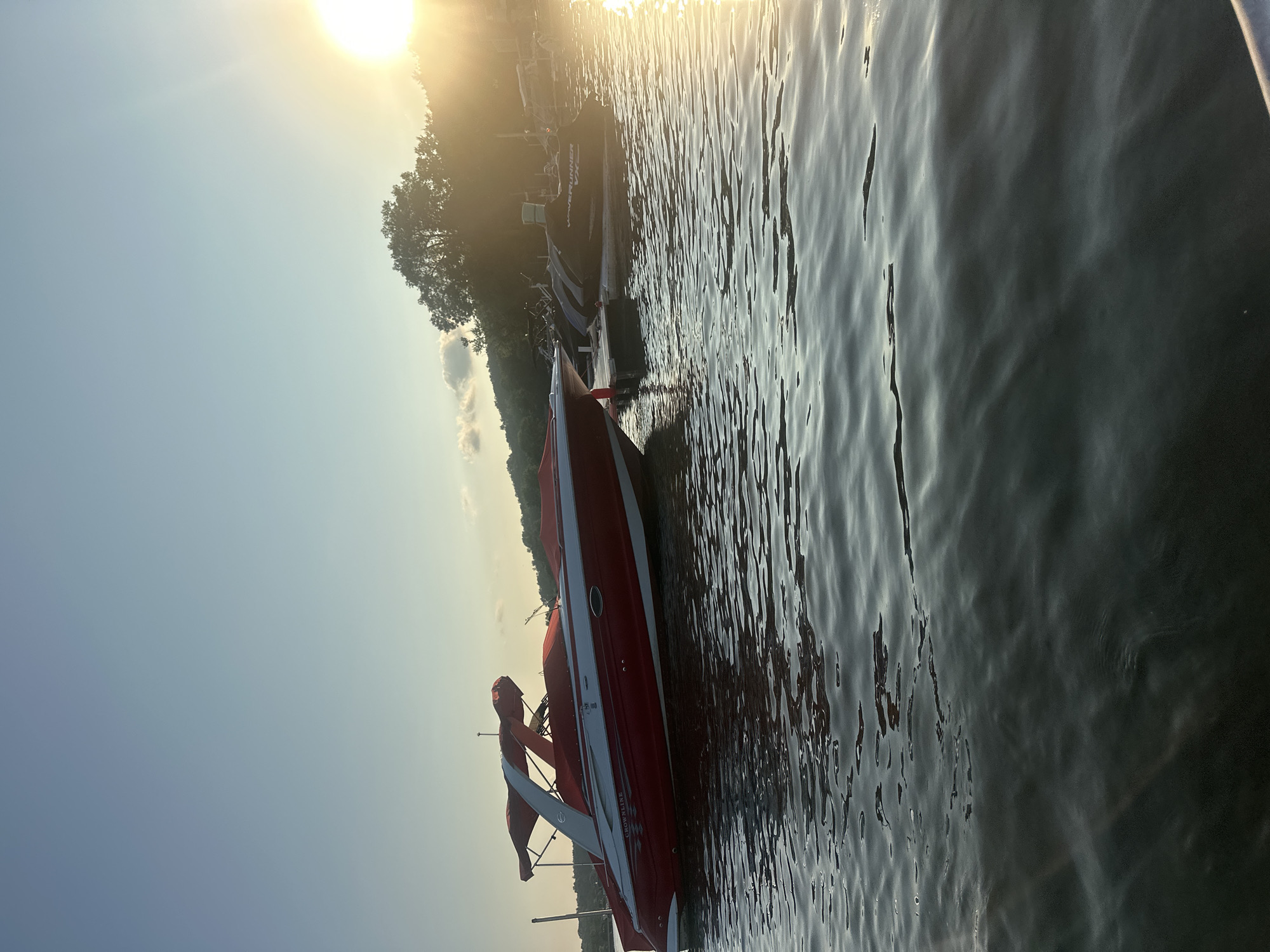 In Algonquin, United States of America, a red and white Crownline boat is docked on a body of water, likely a lake or river, under a clear sky with a warm sun setting in the background. The boat is covered by a red canopy. In the background, there are other boats docked, including jet skis, and a tree-lined shore. The water is calm with gentle ripples, reflecting the sunlight.