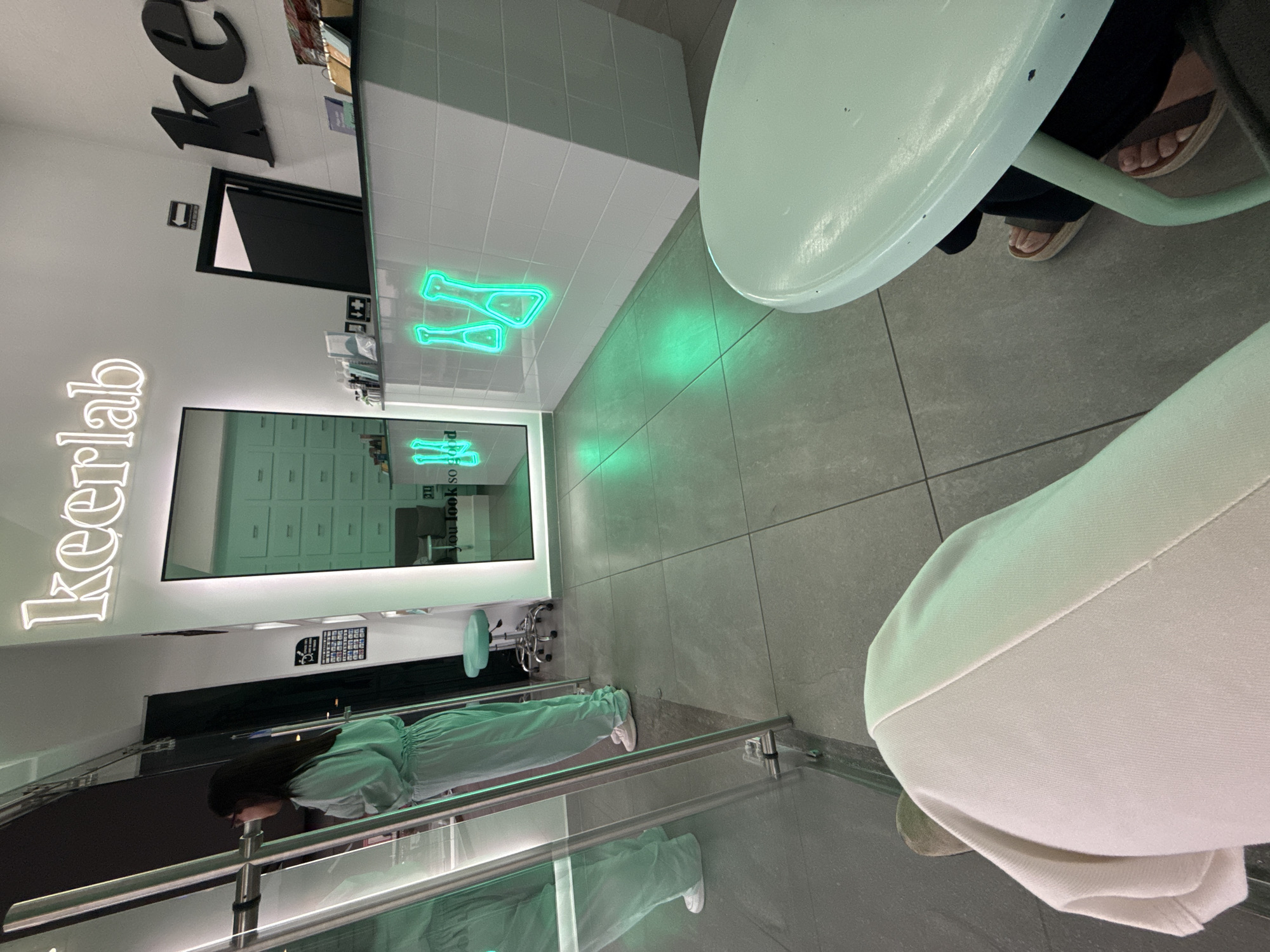 The image shows an interior scene with a person wearing a light green jumpsuit and white sneakers reflected in a glass barrier. A neon sign that reads "keerlab" is visible on the wall. A table with people seated around it is also present, one of whom is wearing sandals. The overall color scheme of the scene is bright, with a focus on white and light green. Two green glowing paddles are seen hanging from the wall. The photo was taken inside of Keerlab at San Bernardino Tlaxcalancingo, Mexico.