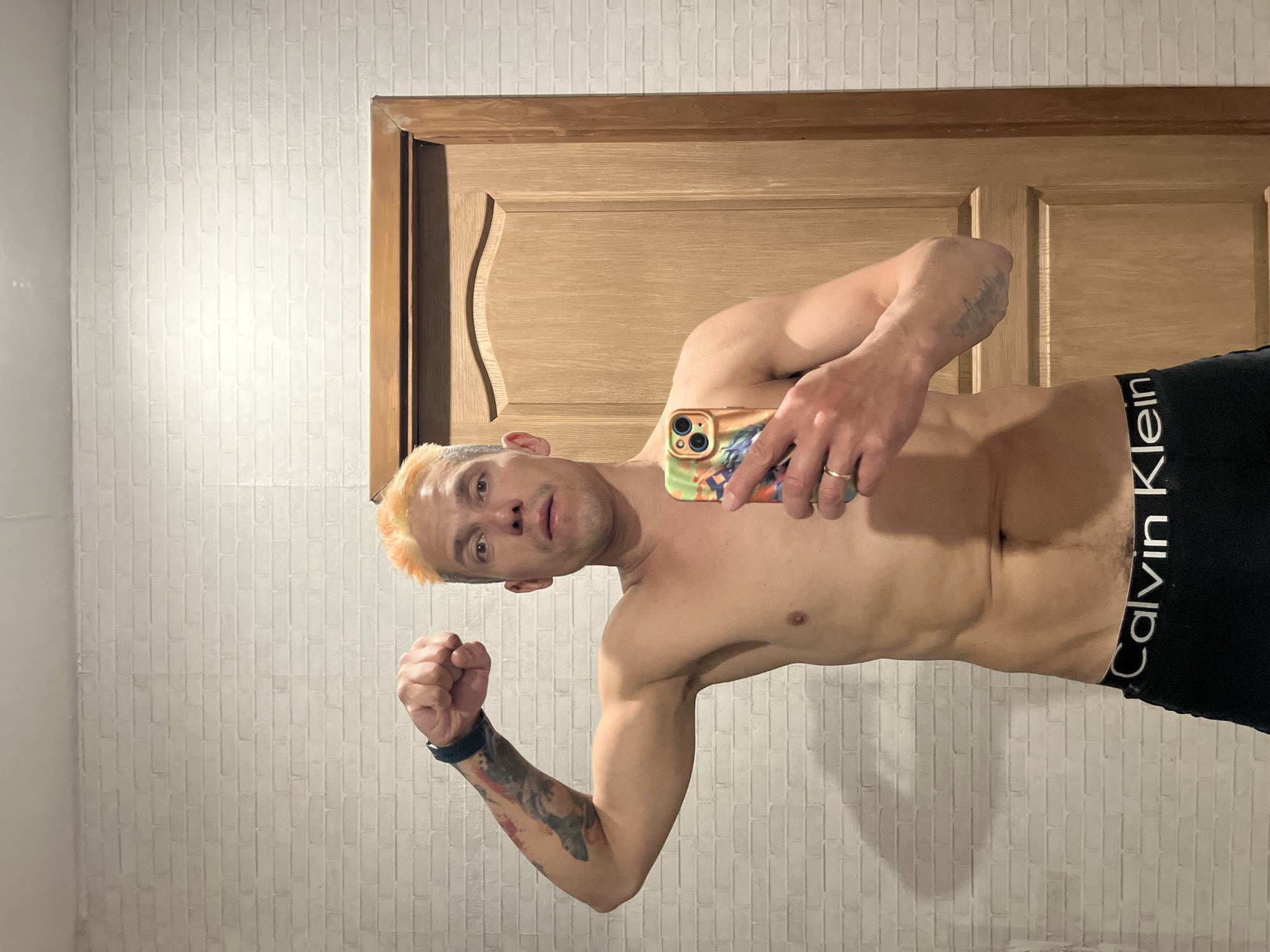 A shirtless man with blonde-dyed hair poses for a selfie indoors. He is wearing black Calvin Klein underwear. He is holding a smartphone. His left arm is flexed, displaying a tattoo. The background features a door and a brick-patterned wall. There is no information to definitively identify the location as the city of Chaikovsky, Russia.