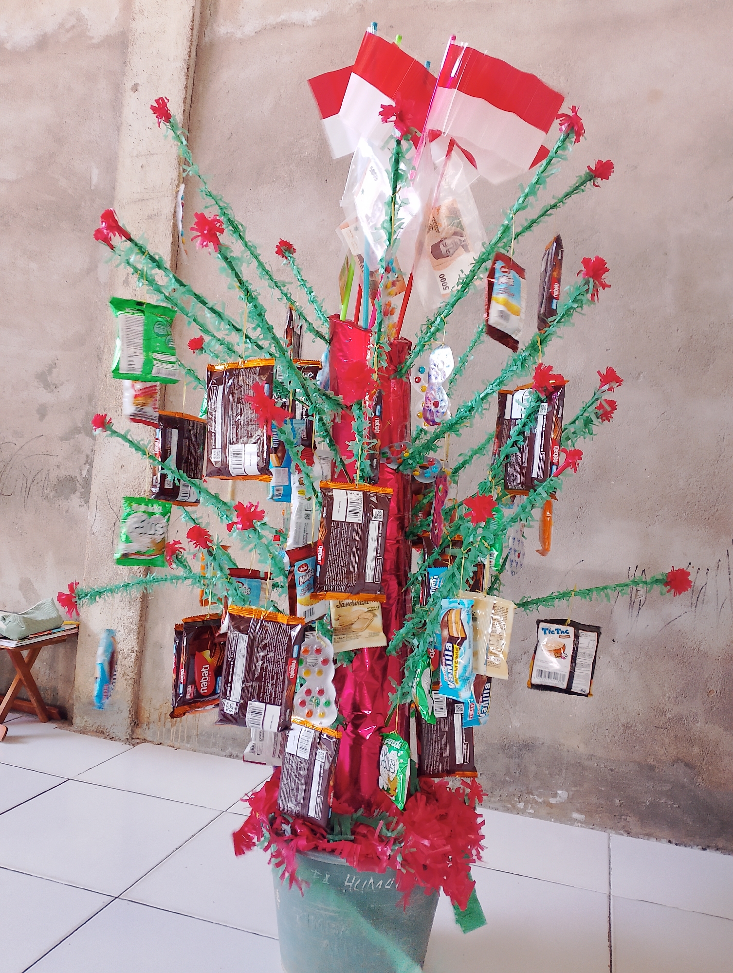 This image captures a decorative tree adorned with Indonesian flags and various packaged snacks and candies. The tree, made of green artificial foliage and red accents, stands in a green bucket labeled with what appears to be "EX HUMA." The backdrop is a textured, light-colored concrete wall, and the floor is tiled with white squares. A small wooden stool with some items on it is visible to the left of the tree. The overall scene suggests a festive or commemorative display, possibly for a local celebration in Bobong, Indonesia.