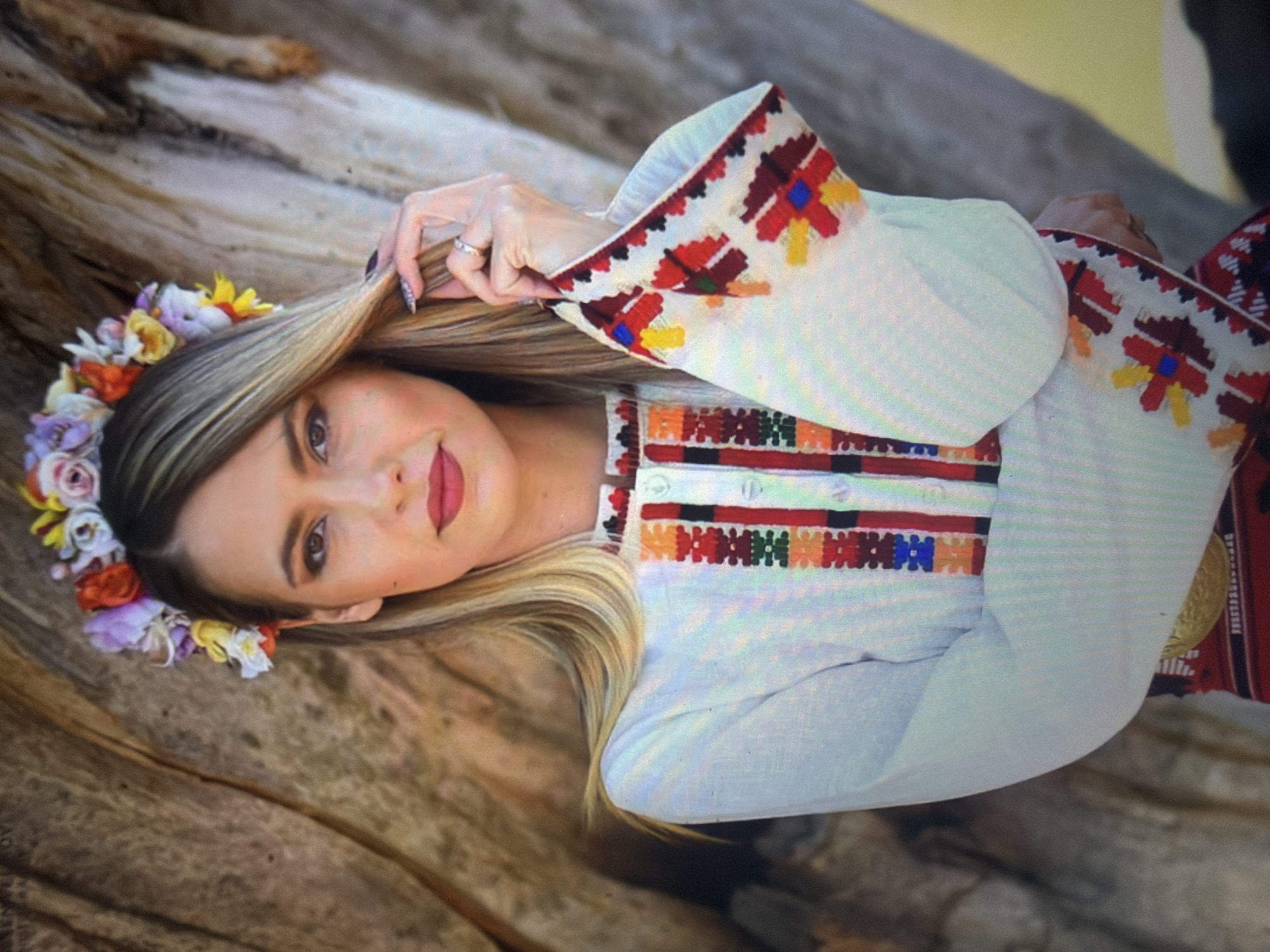 A young adult female is depicted in a frontal, waist-up portrait, positioned outdoors, likely in Shumen, Bulgaria, against the backdrop of a deeply grooved tree trunk. She has long, straight, blonde hair, parted to her right, and wears a vibrant floral wreath or crown on her head, featuring artificial flowers in various colors including red, orange, yellow, purple, pink, and white. She gazes directly at the viewer with a neutral expression; her brown eyes are enhanced with makeup, and her lips are painted red. Her left hand (viewer's right) is raised, gently touching her hair near her cheek; a ring is visible on her left ring finger, and her fingernails are painted a dark color, possibly with glitter.
She is dressed in a traditional-style white long-sleeved garment, possibly a blouse or dress. The garment features intricate geometric embroidery along a central vertical placket and on the wide cuffs of its sleeves. The embroidery on the placket includes patterns in red, orange, black, blue, and green, with white buttons securing the opening. The sleeve cuffs display prominent red, yellow, and blue patterns on a white background. A hint of a red, embroidered lower garment and a gold-toned belt are visible at the bottom edge of the frame. The background is composed of the rough, textured bark of a large tree, displaying natural shades of brown, tan, and grey. The scene is illuminated by natural, bright daylight.