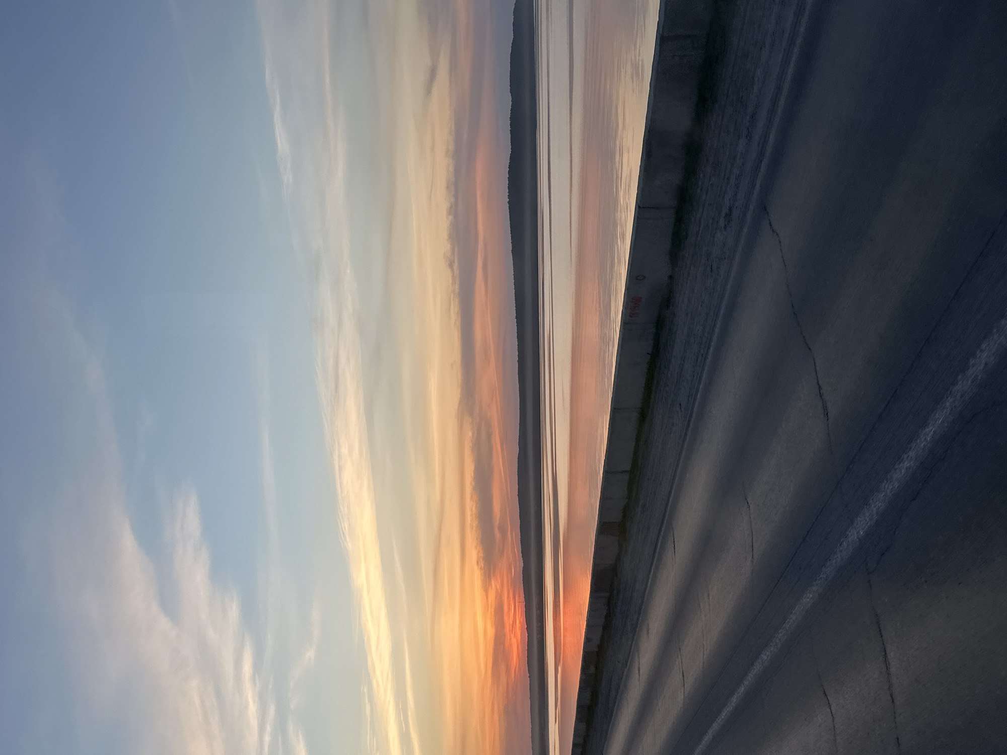 The image depicts a road and sunset in Chaikovsky, Russia. A paved road stretches across the lower right of the frame, its surface cracked and showing lane markings. Adjacent to the road is a concrete barrier. Beyond the barrier, a body of water reflects the colors of the sunset. The sunset is a gradient of blues, oranges, and yellows. A distant treeline is faintly visible between the water and the sky.