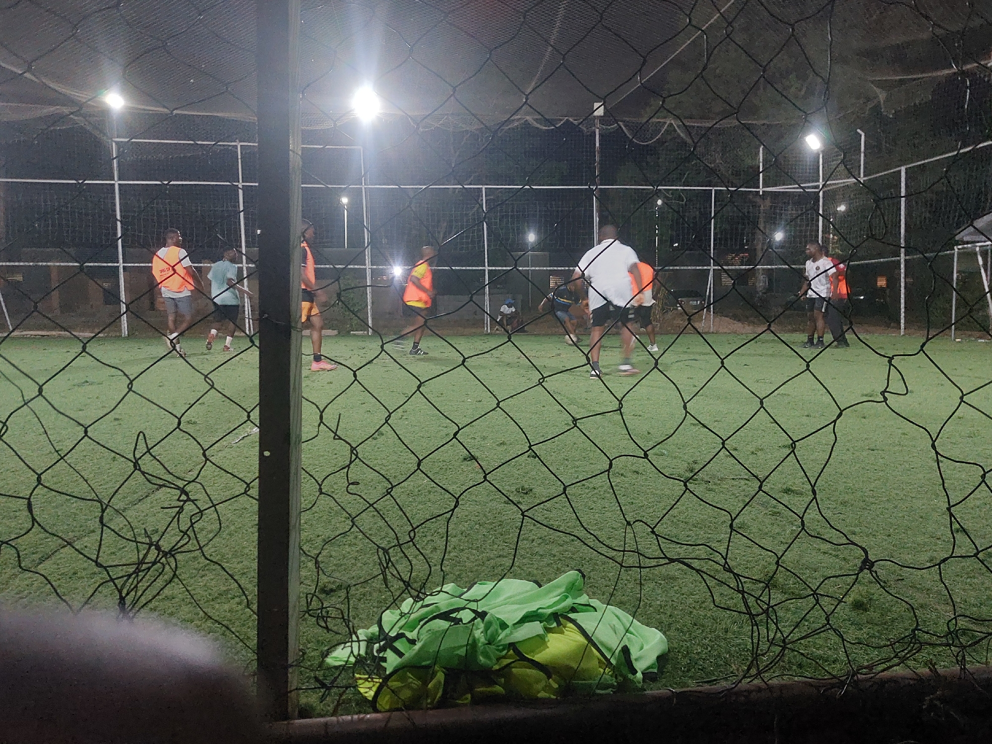 The image depicts a nighttime soccer match in Bar Jirgi Summa, Nigeria, viewed through a black wire mesh fence. Approximately 10-12 adult males are present on a brightly illuminated artificial green turf field. Several players wear athletic shorts and shirts; some are distinguished by bright orange or yellow/lime green pinnies. Multiple bright floodlights on tall poles illuminate the field, casting light from above. In the immediate foreground, a black wire mesh fence, exhibiting some damage and loose strands, obstructs part of the view. A dark metal fence post is visible vertically on the right side of the foreground. On the artificial turf near the fence, a pile of folded green and yellow garments, likely sports vests, is visible. The background consists of dark foliage or trees against the night sky, with additional white structural elements of the field or surrounding facilities. The players are in various dynamic poses, actively engaged in the game across the field.