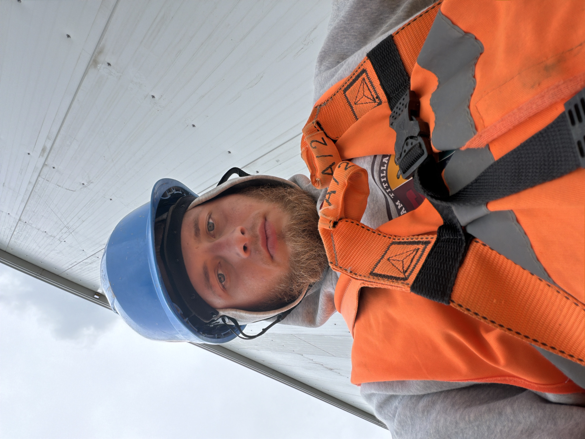 A low-angle, close-up selfie shows a young Caucasian man wearing a blue hard hat, a grey hoodie, and an orange safety vest with black and grey reflective stripes. He has light skin, blue eyes, and a light brown beard. The man is looking directly at the camera. The background is a pale grey, corrugated metal wall, suggesting an industrial or construction setting. The sky above is overcast. The man is wearing a safety harness with orange straps that cross over his chest and a black buckle. The location of this image is in Stróża, Poland.