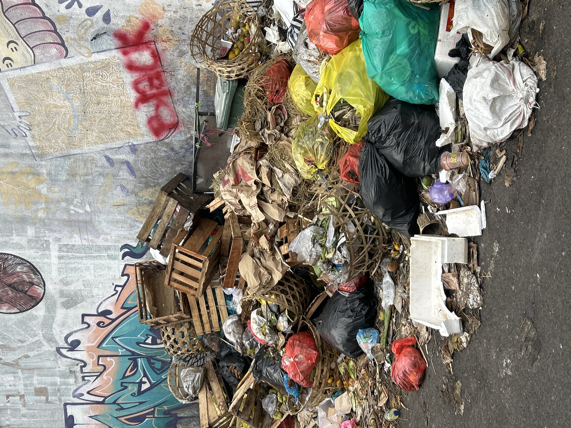 The image depicts a scene of accumulated garbage on an asphalt surface, with a graffiti-covered wall in the background. The garbage consists of various items, including plastic bags filled with waste, wooden crates, baskets, paper packaging, and organic refuse such as fruits and straw. The wall behind the debris is adorned with colorful graffiti, some of which appears to be spray-painted tags and artwork. In the upper right corner of the wall, the word "BLACK" is spray-painted in red. The overall impression is one of neglect and unsanitary conditions. The location is identified as Denpasar, Indonesia, suggesting an urban environment where waste management may be a challenge. There are no people visible in the image. The lighting suggests it could be daytime.