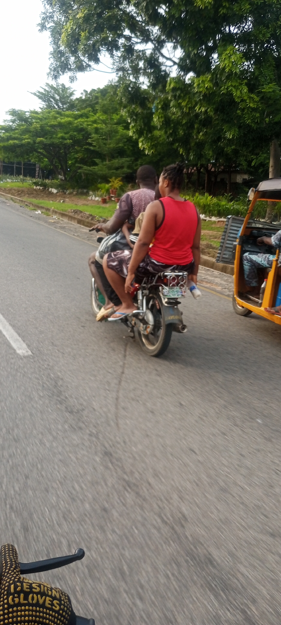Two individuals are riding a motorcycle on a paved road in Lokoja, Nigeria. The person in the rear, a woman with braided hair, is wearing a red tank top and dark shorts. The person in front, likely a man, is wearing a patterned short-sleeved shirt and dark shorts. They are traveling on a road with a white painted line marking the lane. To the right of the motorcycle, a yellow three-wheeled auto-rickshaw is visible. The environment includes lush green trees and foliage along the side of the road, with some potted plants and fencing. The sky is overcast. The image is taken from a slightly elevated perspective, possibly from another vehicle.