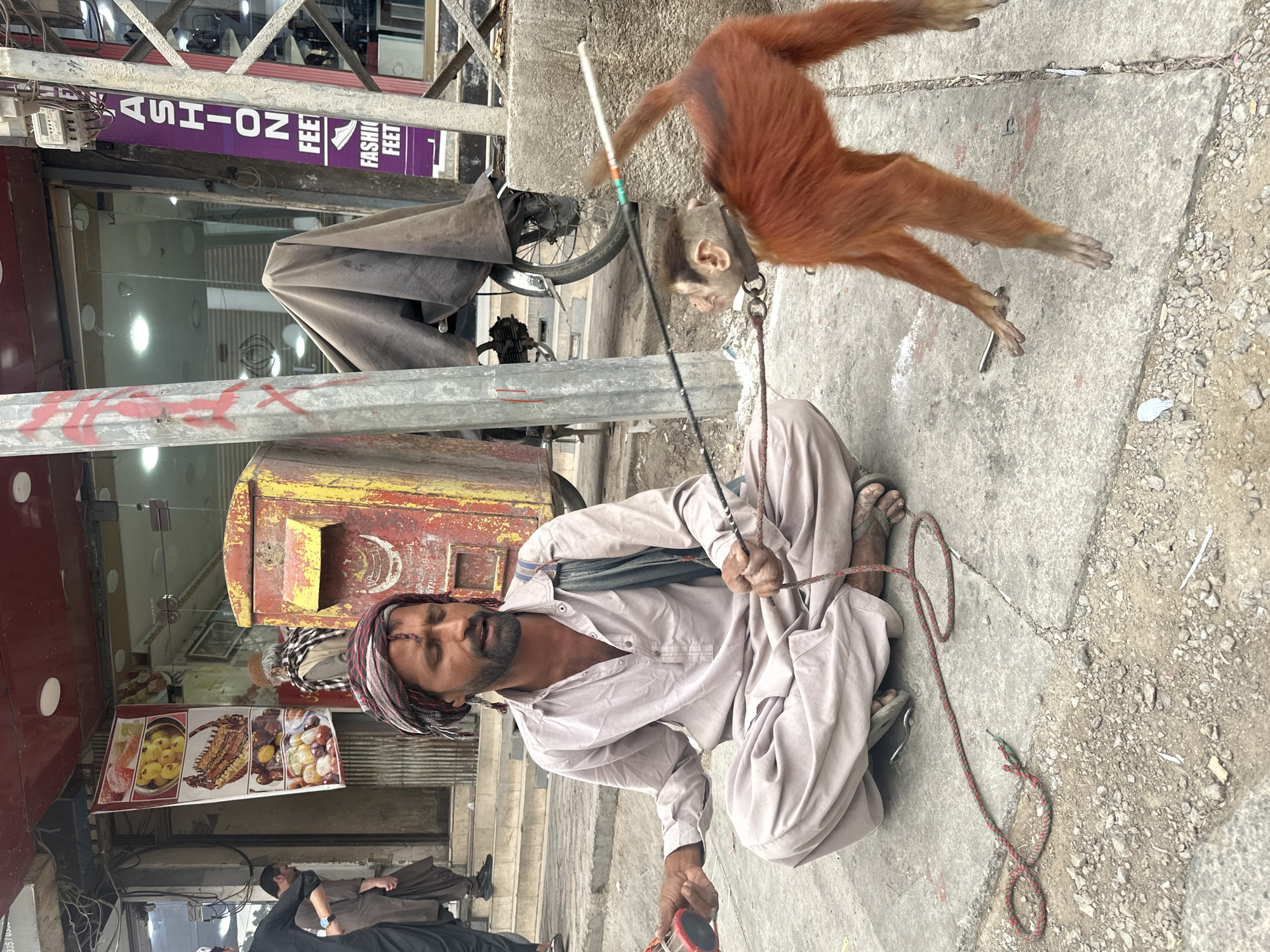 A street scene in Quetta City Tehsil, Pakistan features a man in a light-colored traditional shalwar kameez and a patterned headwrap seated on the ground. He holds a rope attached to a monkey with orange fur that is airborne. A red and yellow post box is to the left of the man. Behind the man is a shop front with a food advertisement. A sign for "Fashion Feet" is visible above the shop. Other people are walking in the background.