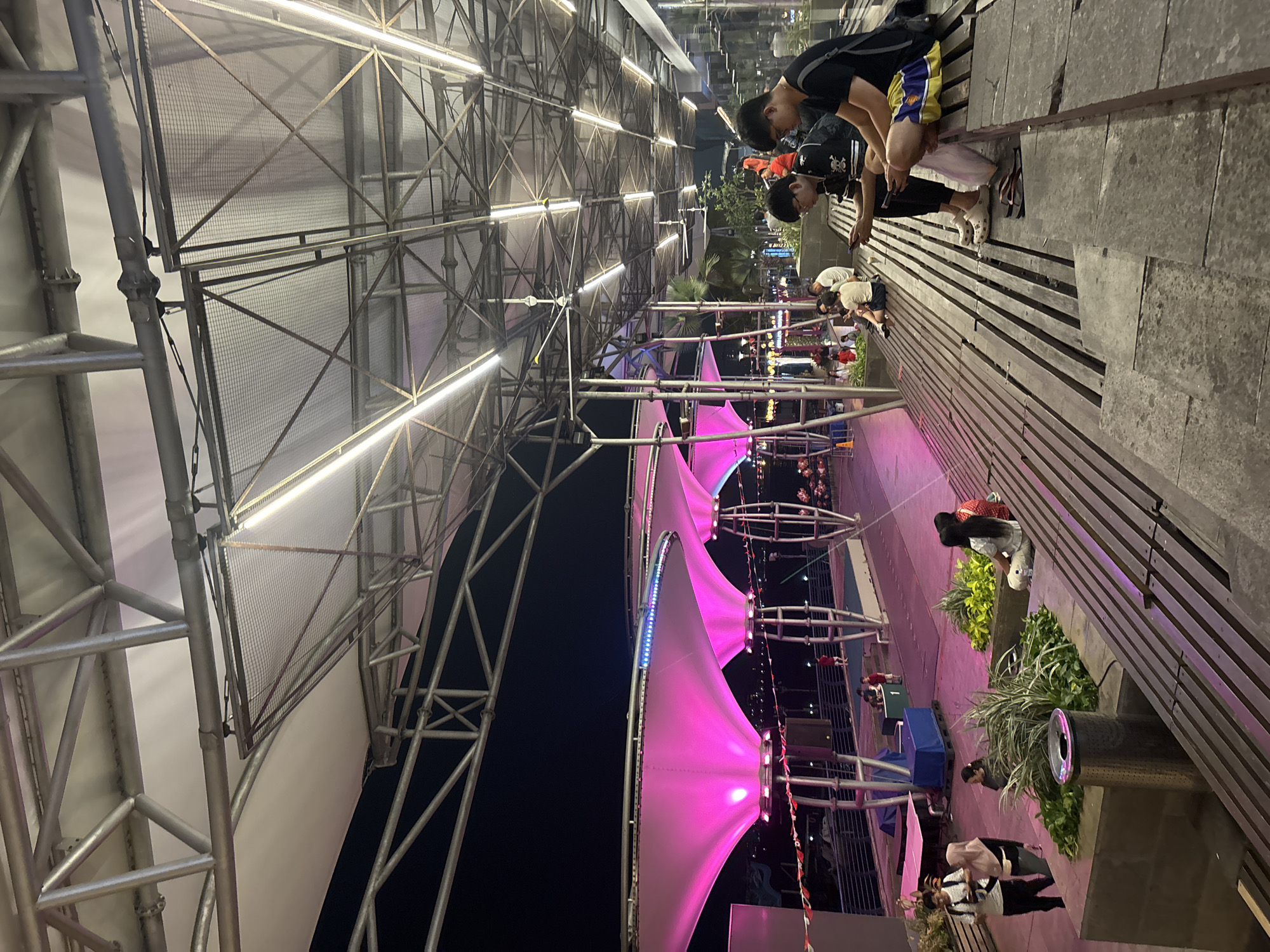 In North Jakarta, Indonesia, at night, an outdoor waterfront area is illuminated by pink-hued fabric canopies supported by metal structures. A group of people are visible, some seated on wooden benches and stone steps, others standing and walking. Two young men are in the foreground, seated on steps, both engrossed in their mobile phones. One man wears a black t-shirt with a white graphic and striped shorts, while the other wears a black t-shirt and shorts. A woman in a white shirt and patterned pants sits beside them, also looking at her phone. Further in the background, more individuals are scattered throughout the walkway, some interacting with each other. Lush green plants are integrated into the seating area, and overhead, a complex metal framework supports white fabric panels and numerous fluorescent lights. The scene appears to be a public promenade or entertainment venue by the water.