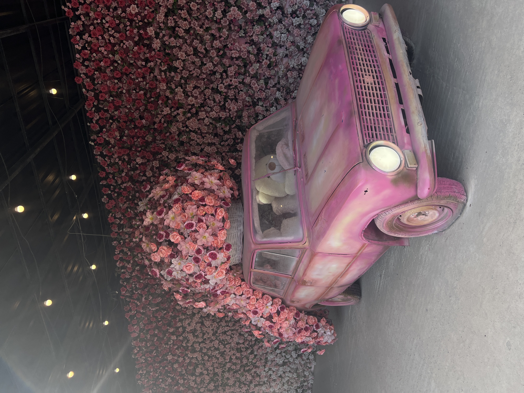 A pink vintage car is parked in front of a wall covered in pink and red artificial flowers. The car is decorated with a large basket overflowing with pink and white artificial flowers placed on its roof. Inside the car, two large white teddy bears are visible. The scene is illuminated by strings of warm-toned lights hanging from a dark ceiling above. The ground appears to be a concrete surface. The car is positioned to the right of the frame, with the flower wall occupying the majority of the background. This setup is located in Ternopil, Ukraine.