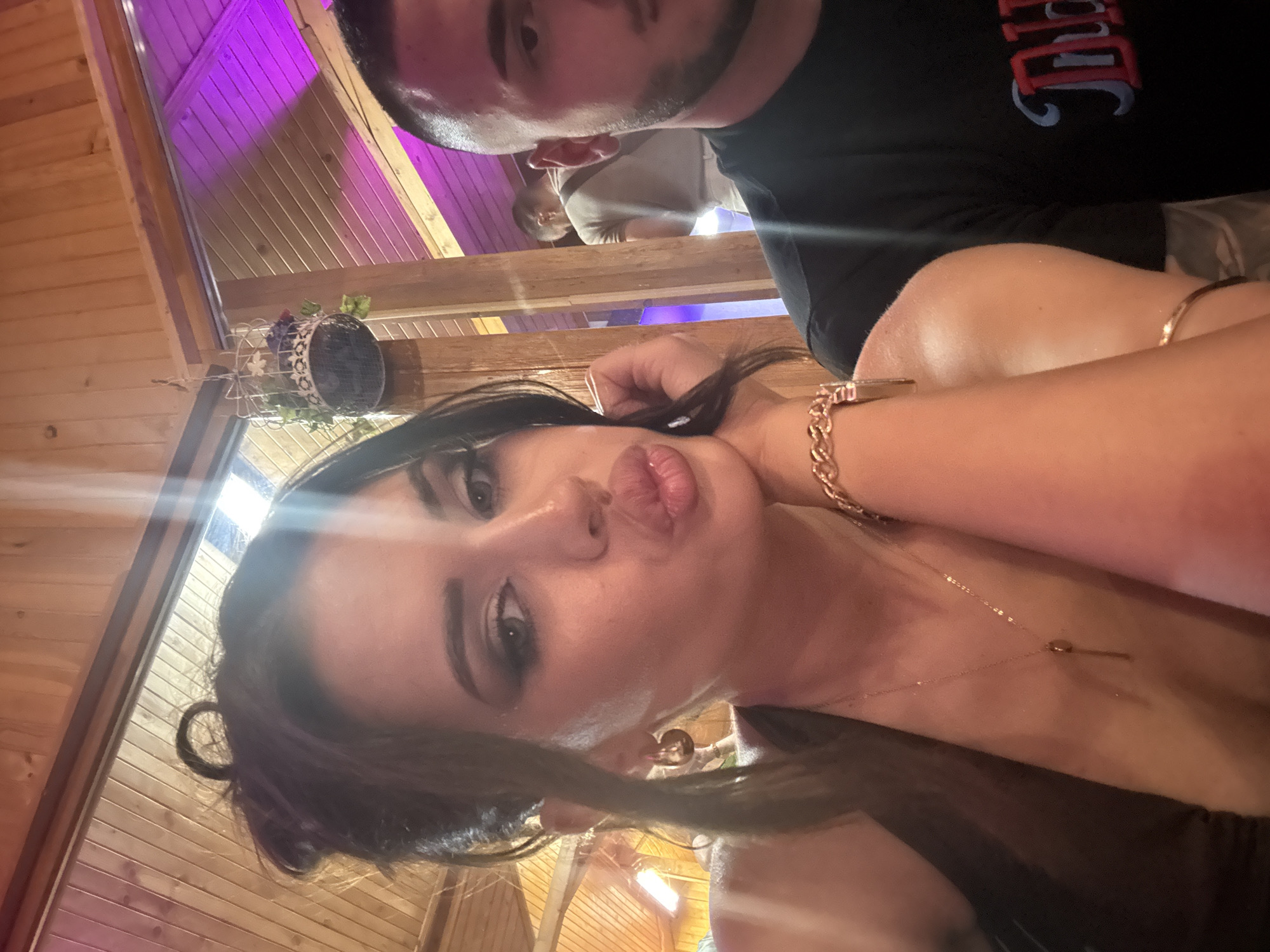 This image captures an indoor scene, likely a restaurant or bar, located in Debrashtitsa, Bulgaria. The foreground is dominated by a young woman posing for the camera, occupying the left and center of the frame. She has dark hair styled in a messy bun with a distinct loop on top and some loose strands framing her face. Her eyes are heavily made up with dark shadow and liner, and her eyebrows are well-defined. She is making a puckered, pouting expression with full lips, looking directly into the camera. She wears gold-toned hoop earrings, a delicate gold chain necklace with a small, elongated pendant (resembling a key or bar), a thick gold-toned chain-link bracelet on her right wrist, and a thin gold bangle on her upper right arm. Her right hand is raised, with fingers gently touching her neck/chin area. She appears to be wearing a dark, possibly brown or black, sleeveless or halter-style top.
To her right, partially visible, is a young man. Only the right side of his face and upper torso are visible. He has short, dark hair with a faded cut on the sides and a short, dark beard. He is wearing a black t-shirt with a design on the chest featuring red and blue lettering, part of which reads "DID". His gaze is directed slightly downwards and away from the camera.
The background features light-toned wooden paneling on the walls and a sloping wooden ceiling. Various light sources illuminate the scene: a prominent bright light flare (possibly from a window or strong artificial light) is visible at the top center, casting a strong glare. On the left side of the background, a horizontal artificial light source is visible. On the far right, parts of the wooden ceiling structure are illuminated with a distinct purple/magenta light. A small, decorative wire cage with artificial greenery and flowers is suspended from the wooden ceiling towards the upper left background. Further in the background, behind the man, another individual wearing a light-colored t-shirt and trousers is faintly visible, standing.
